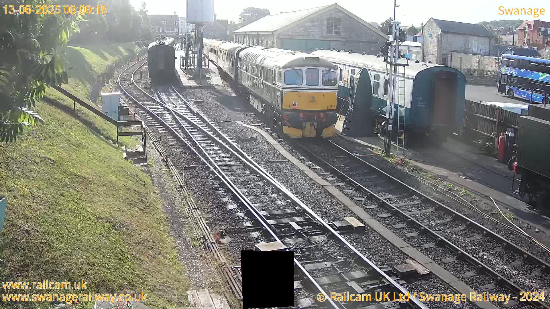 A diesel locomotive leads a passenger train at a railway station on a sunny day.