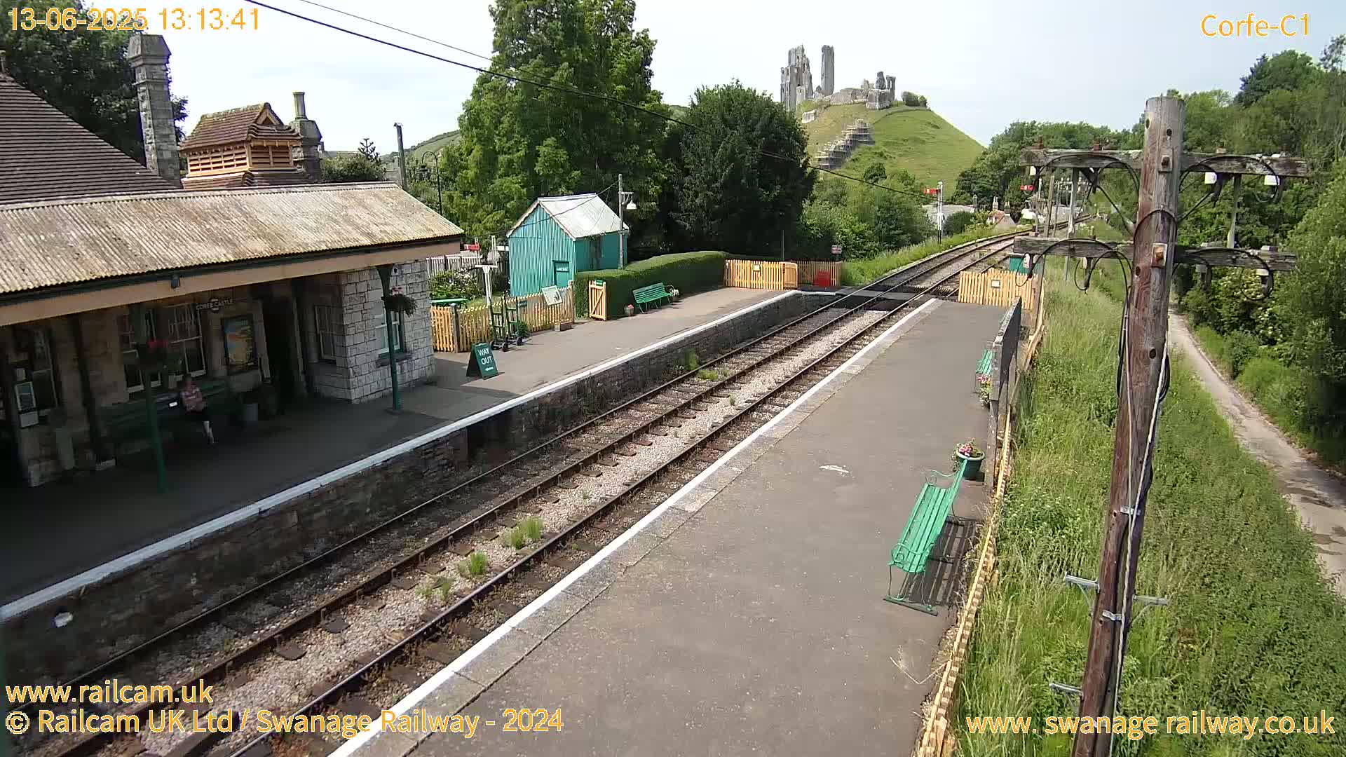 A train station platform with a stone building and a green waiting shelter, overlooking a hill with a ruined castle under a partly sunny sky.