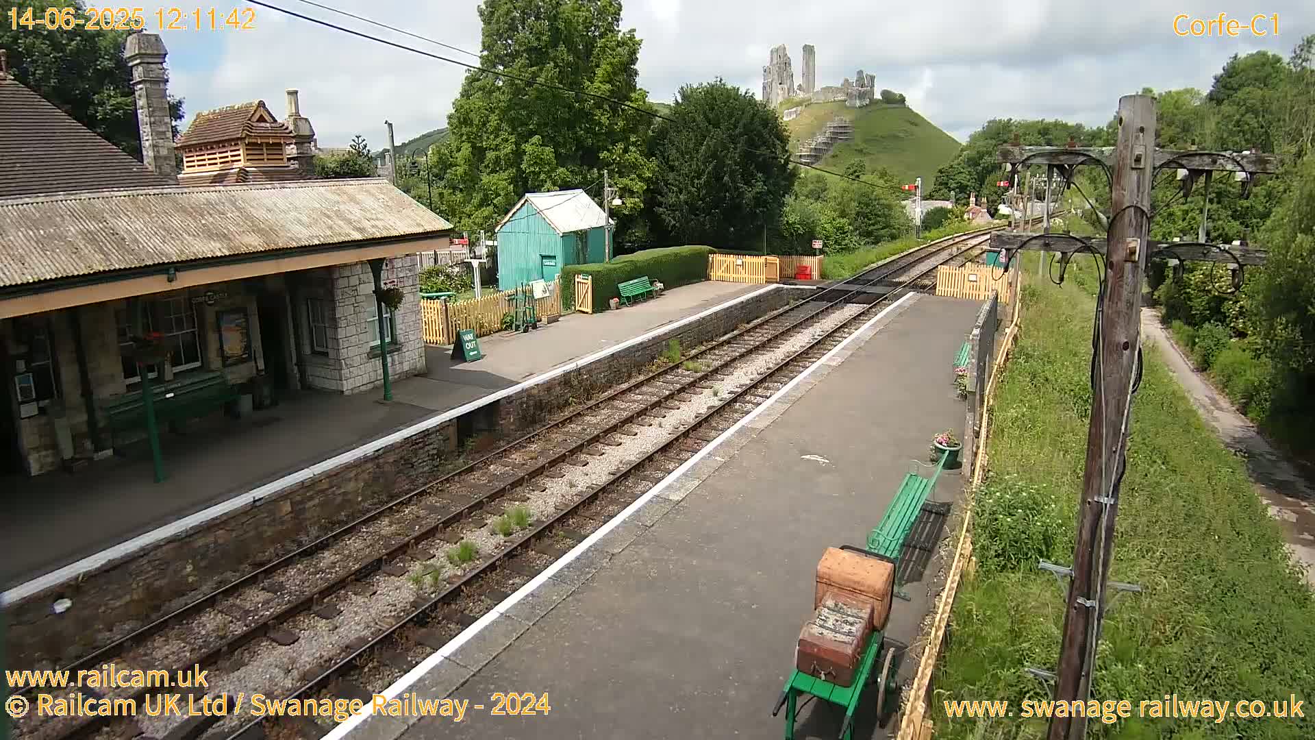 A railway station platform with a stone building, a small green shed, and train tracks, all under a partly cloudy sky with a ruined castle visible on a hill in the background.