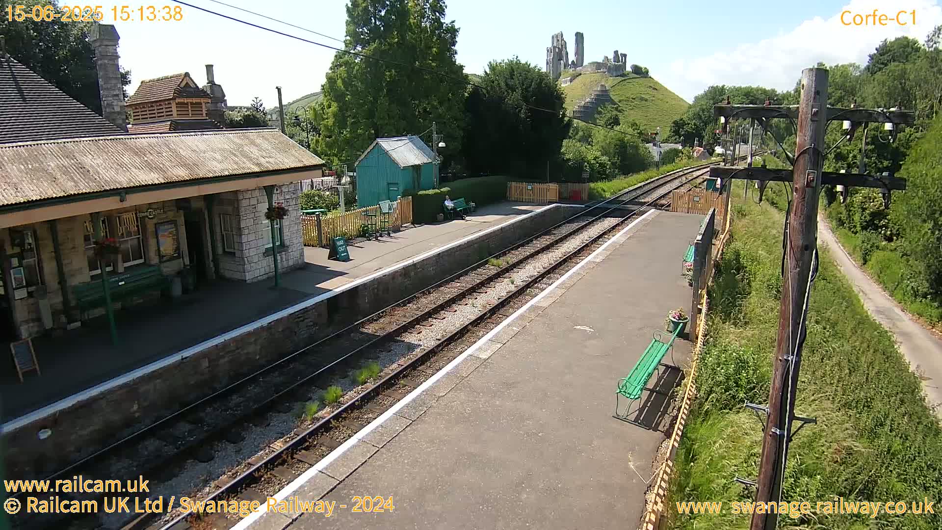 A train station platform with a stone building, a small green shed, and a distant castle ruin visible on a sunny day.
