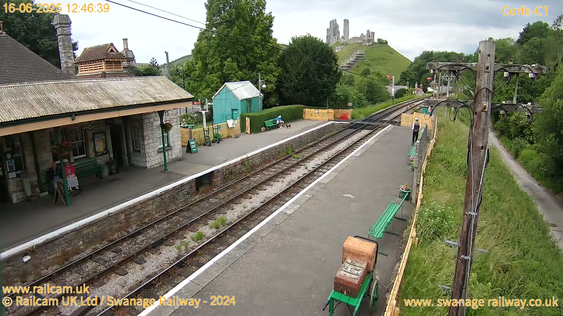 A train station with a stone building, a small green shed, and railway tracks under an overcast sky, with a hilltop castle in the background.