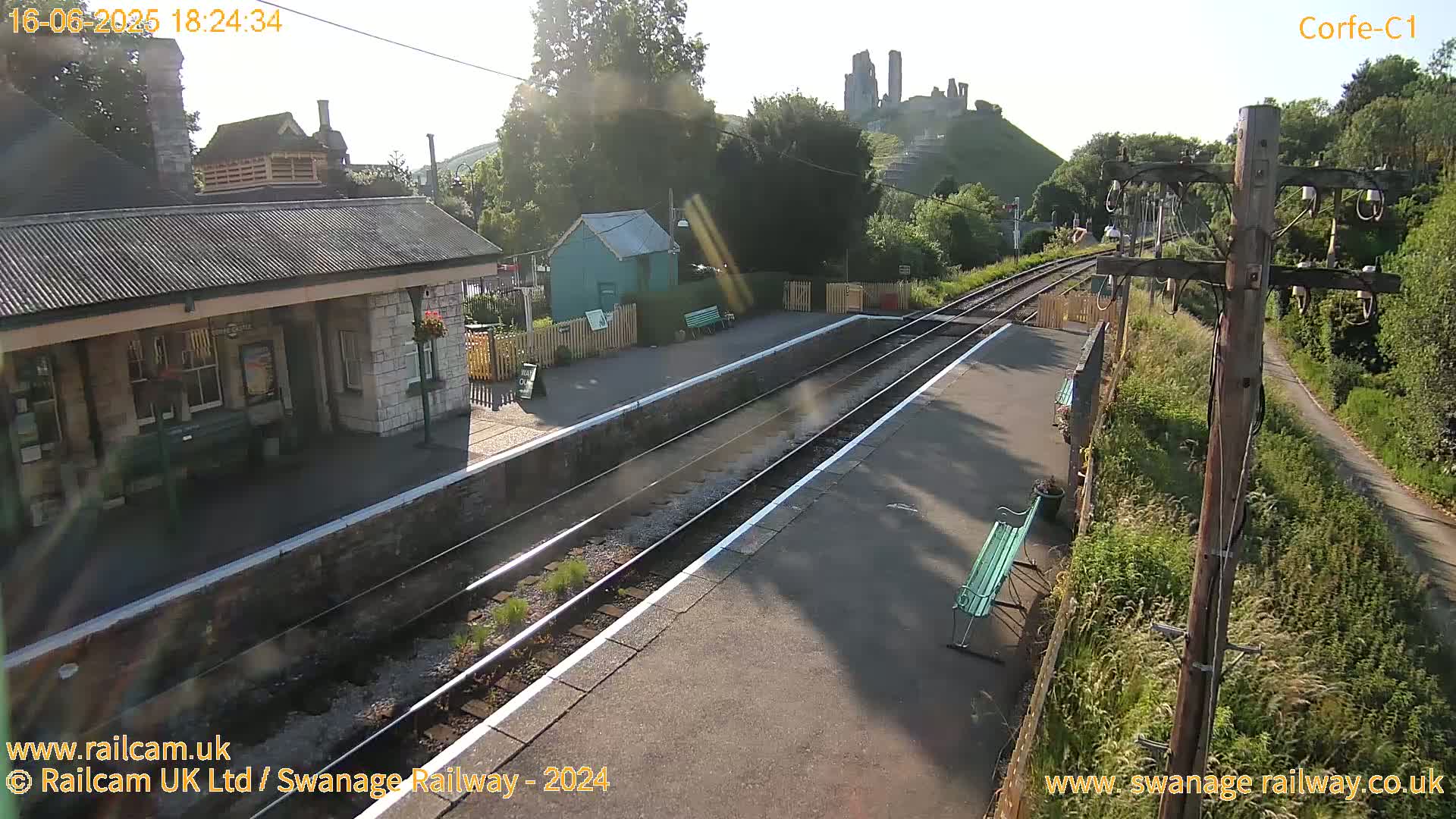 A sunny view of a train station platform with a stone building, a small green shed, and a castle ruin visible in the distance.