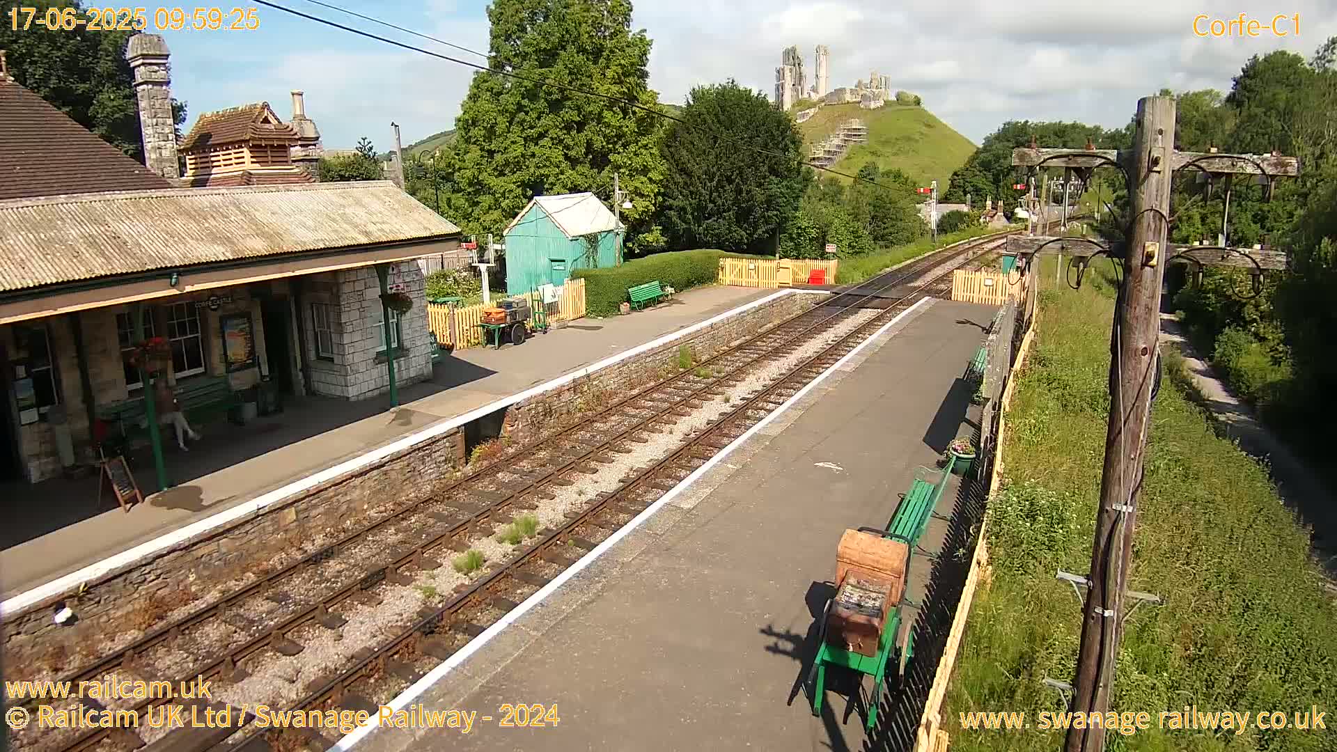 A train station platform with a stone building, a small green shed, and train tracks stretches toward a hilltop with the ruins of a castle under a partly sunny sky.