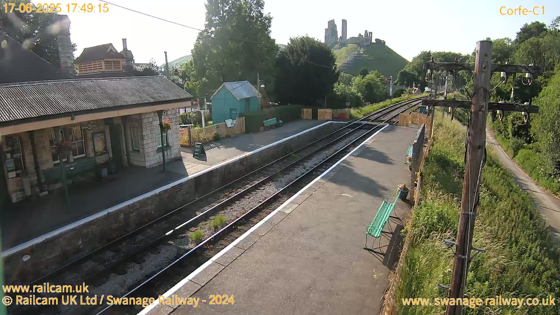 A train station platform with two tracks, a stone building, a small green shed, and a hill with ruins in the background on a sunny day.