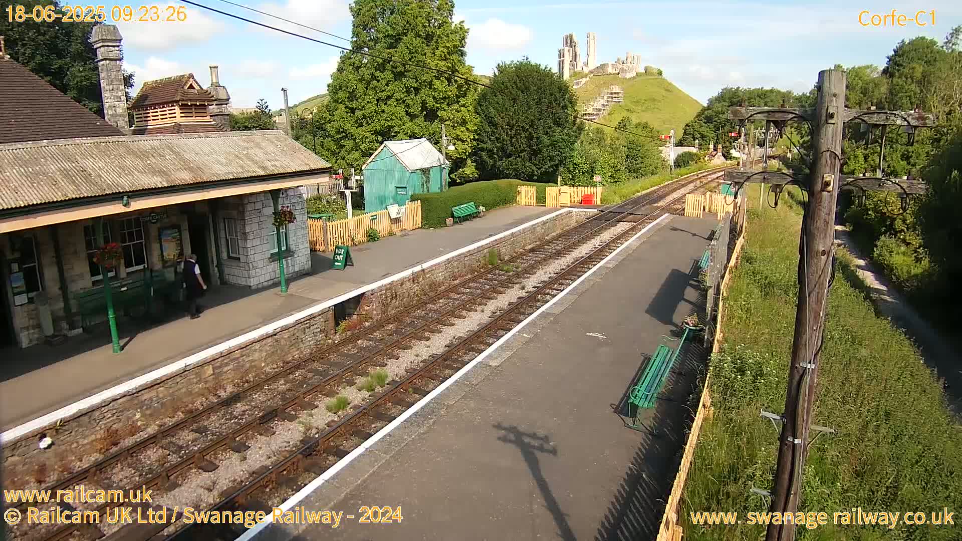 A train station platform with a stone building, a small green shed, and a hill with a ruined castle in the background on a sunny day.