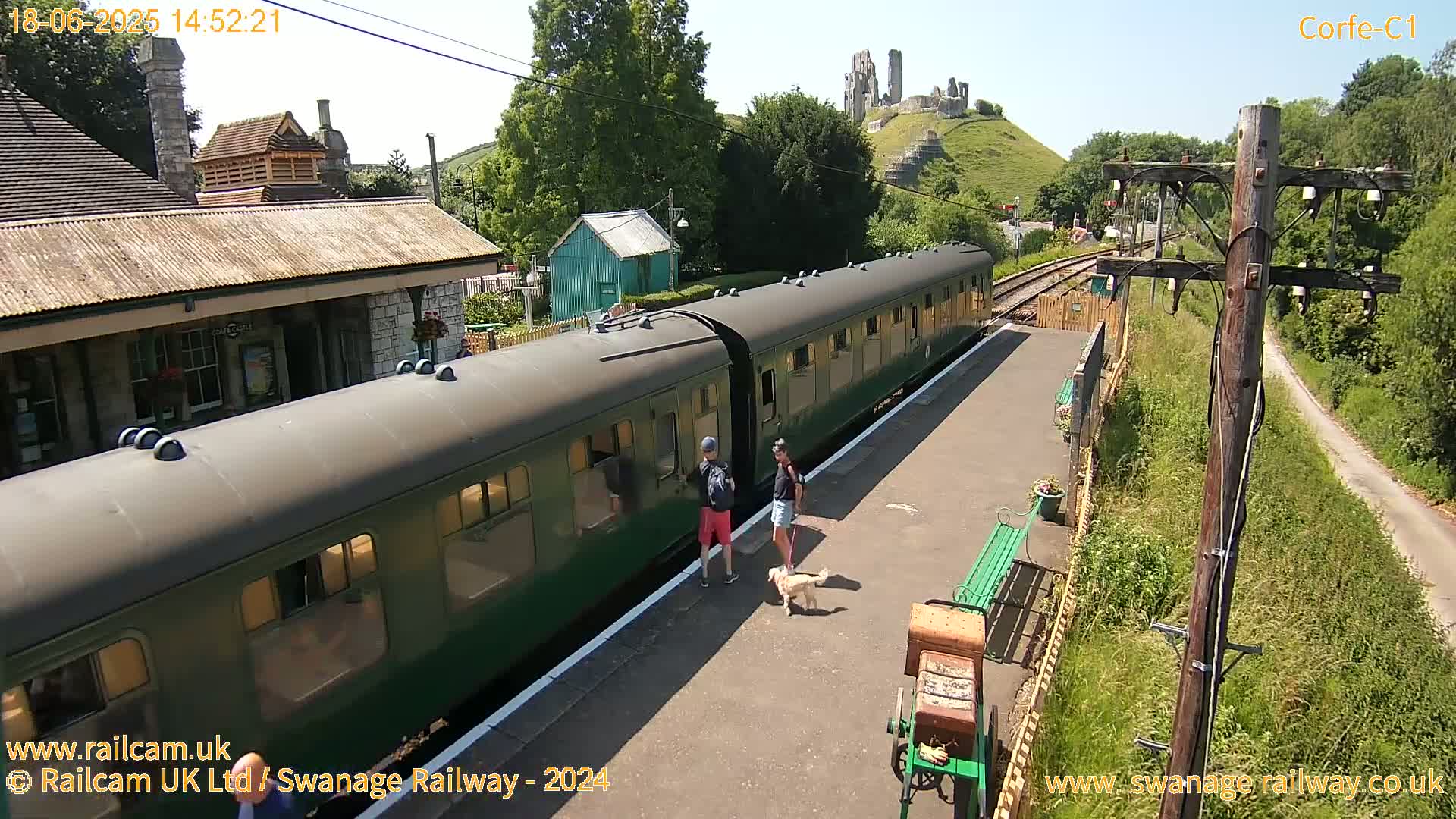 A green passenger train is stopped at a station platform on a sunny day, with a couple and their dog nearby, and a hilltop castle visible in the background.