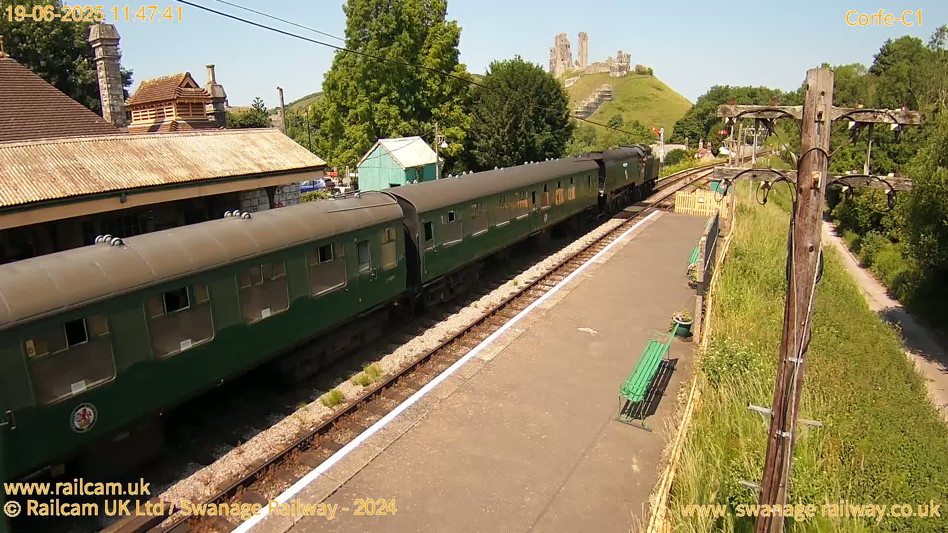 A long green passenger train sits at a railway station on a sunny day, with a ruined castle visible on a hill in the background.