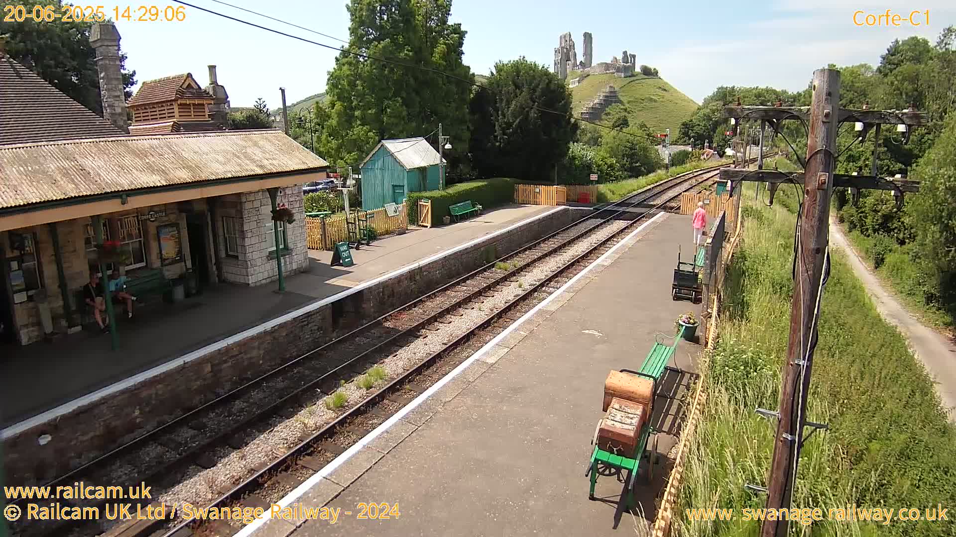 A sunny day at a train station with a stone building, a green shed, and a distant castle ruin visible on a hill.