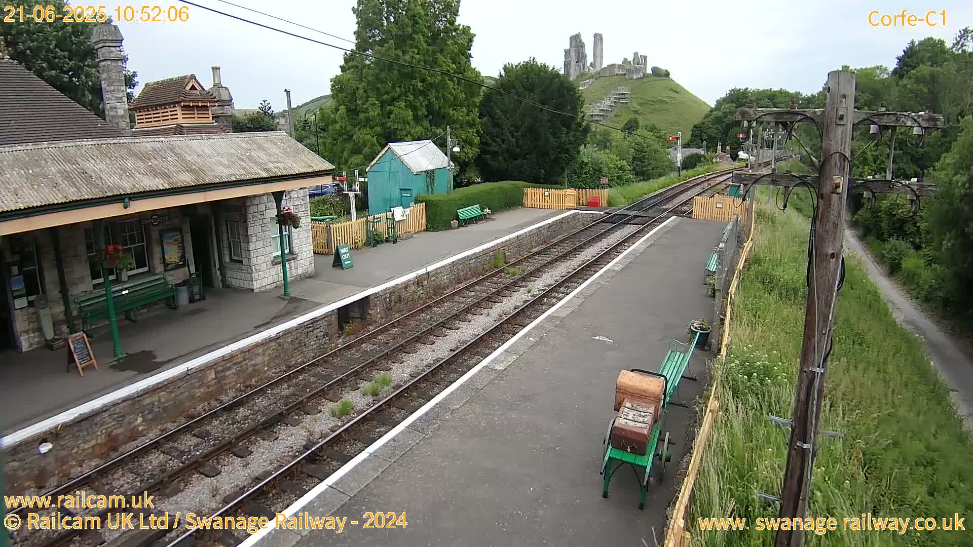 A train station platform with a stone building, a small green shed, and a hilltop castle in the background under partly cloudy skies.