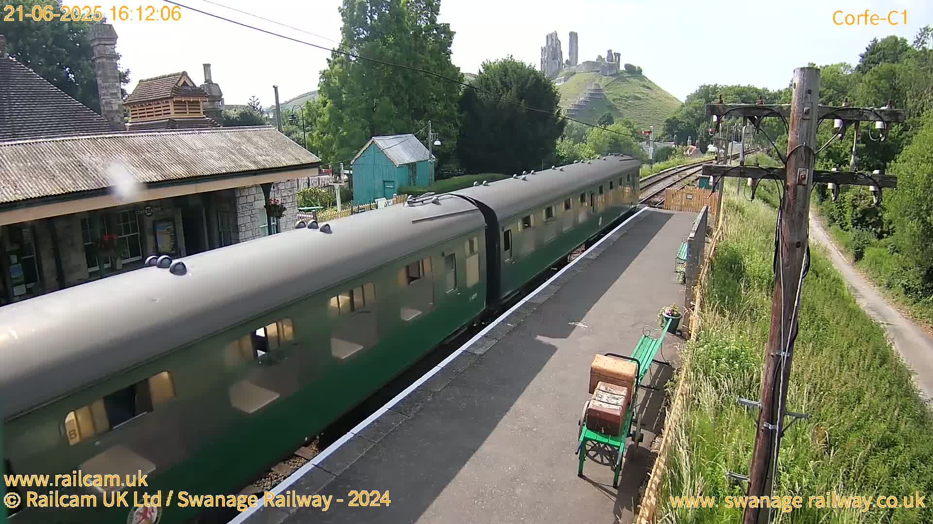A green passenger train sits at a station platform on a sunny day, with a castle visible on a hill in the background.