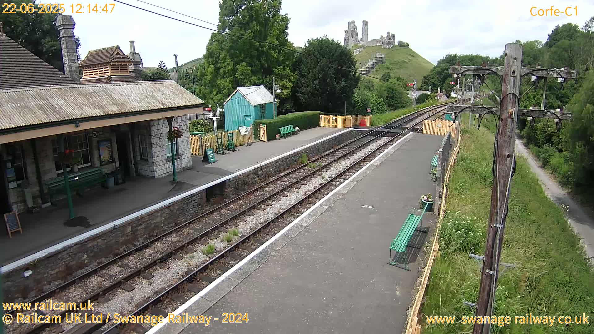 A train station platform with two tracks, a stone building, and a small teal shed, overlooks a hill topped with a partially ruined stone structure under a mostly cloudy sky.