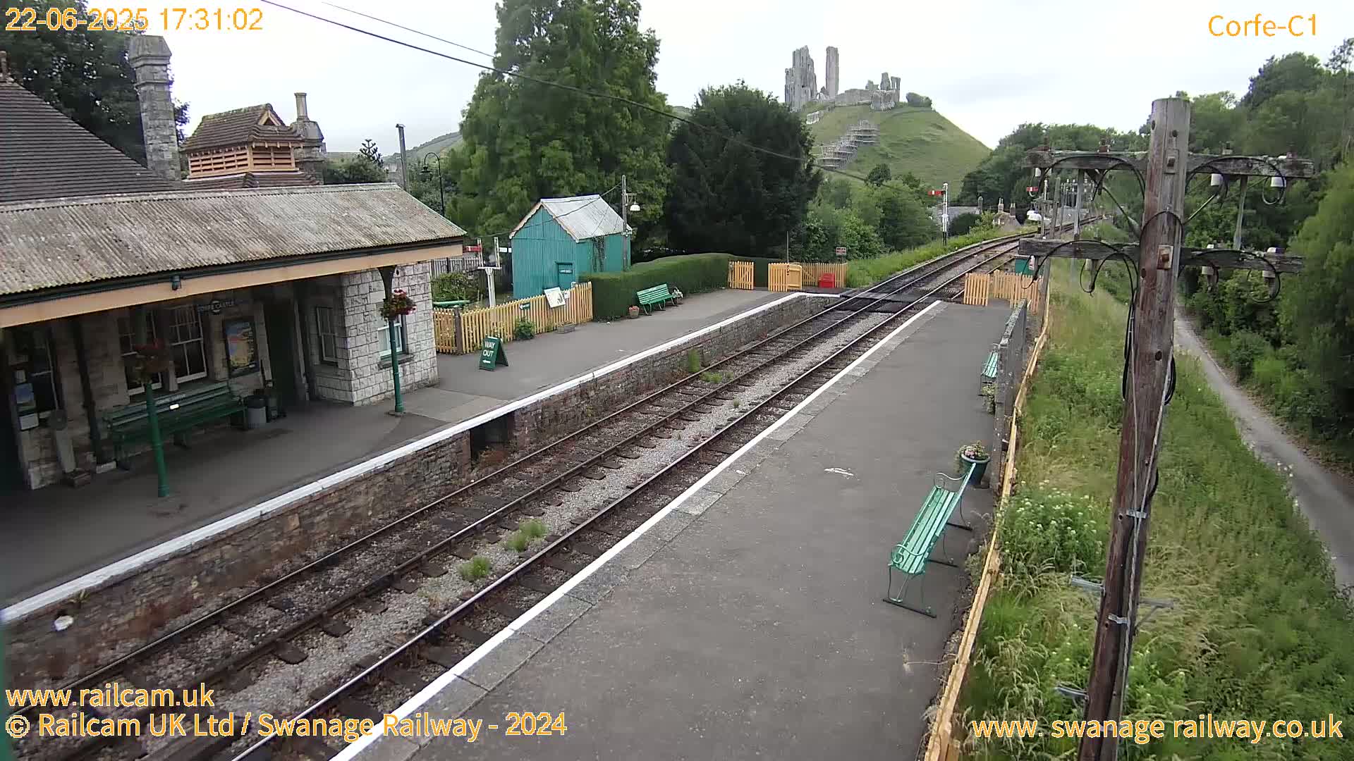 A train station platform with two tracks, a stone building, a small green shed, and a hill with a castle ruin in the background under an overcast sky.