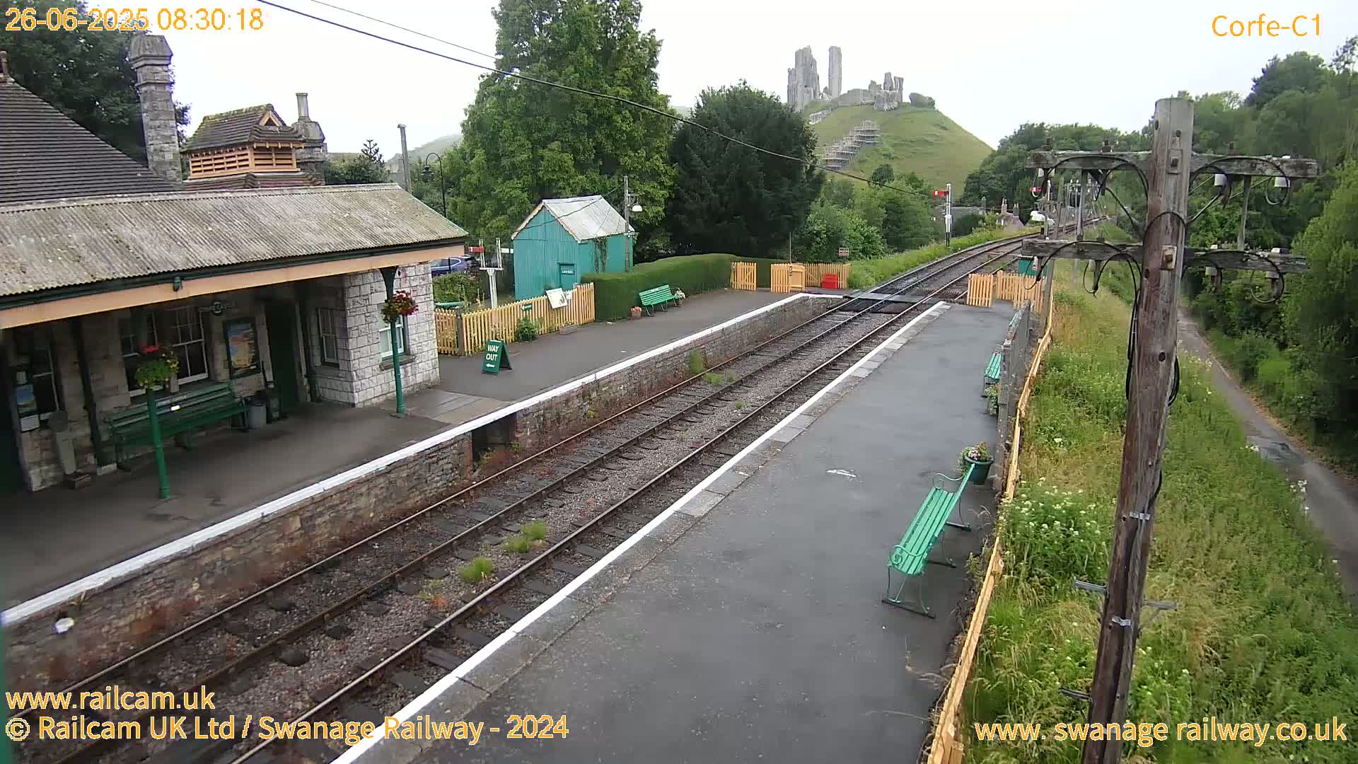 A train station platform with two sets of tracks, a stone building, and a distant hilltop castle is visible under an overcast sky.