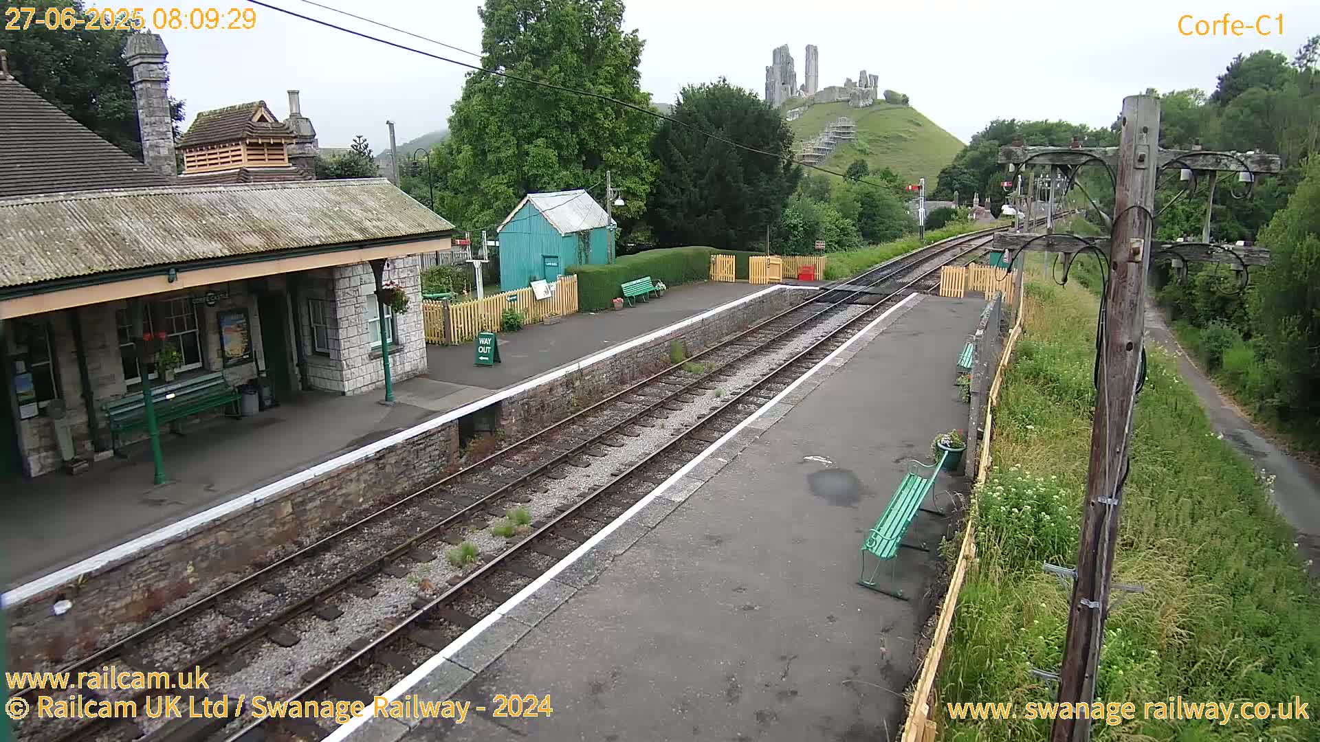 A train station platform with a stone building and benches, overlooking a hill with a ruined castle in the background under an overcast sky.