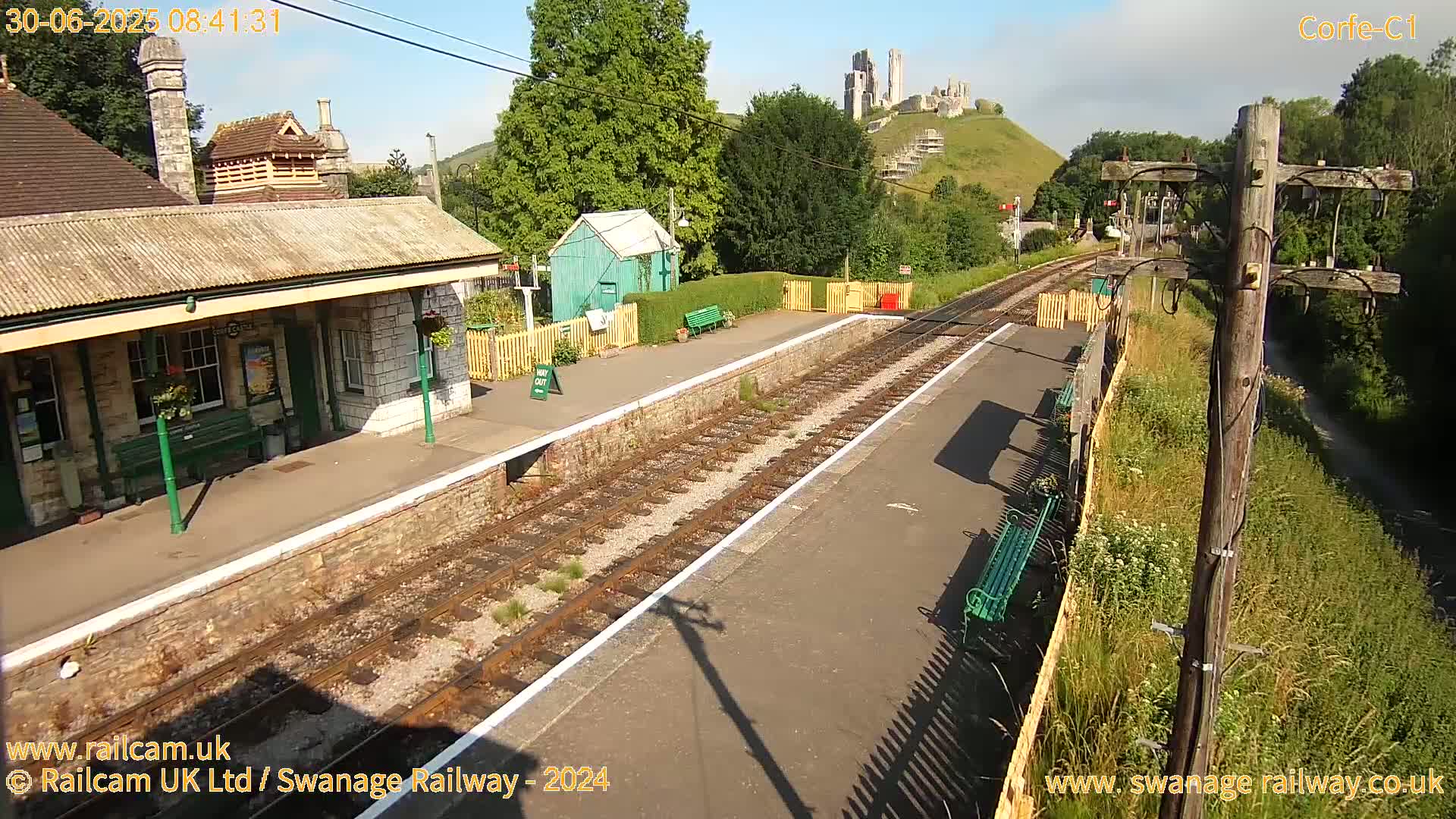 A train station platform with a stone building and a view of a castle atop a hill on a sunny day.