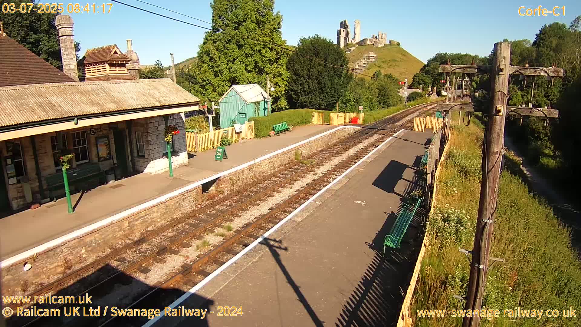 A train station platform with a stone building, a small green shed, and a distant hilltop castle is visible on a sunny day.
