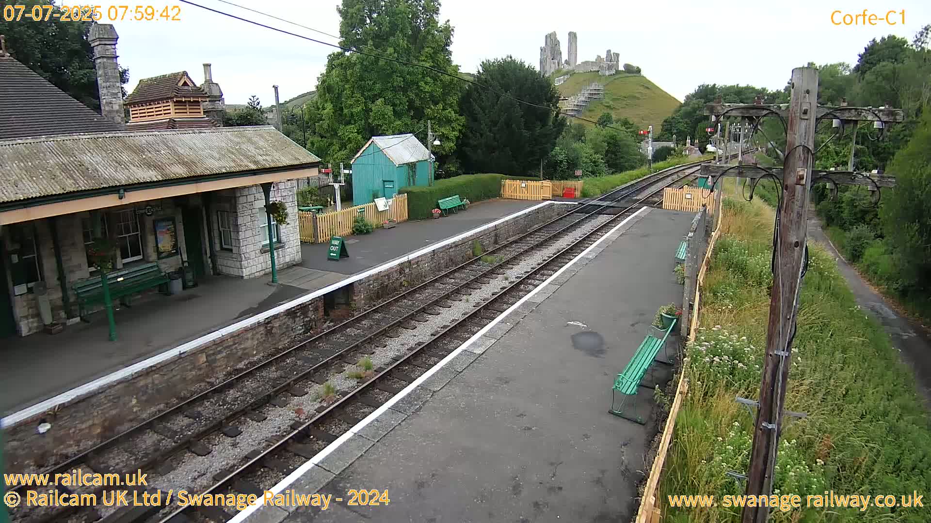 A train station platform with a stone building, a small green shed, and train tracks, all under an overcast sky, with a hilltop castle visible in the background.