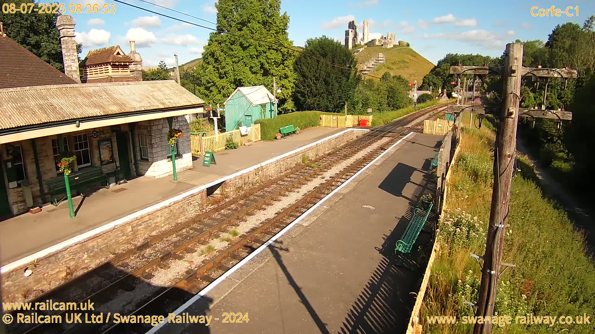 A train station platform with a stone building and a castle visible on a hill in the background under a partly sunny sky.