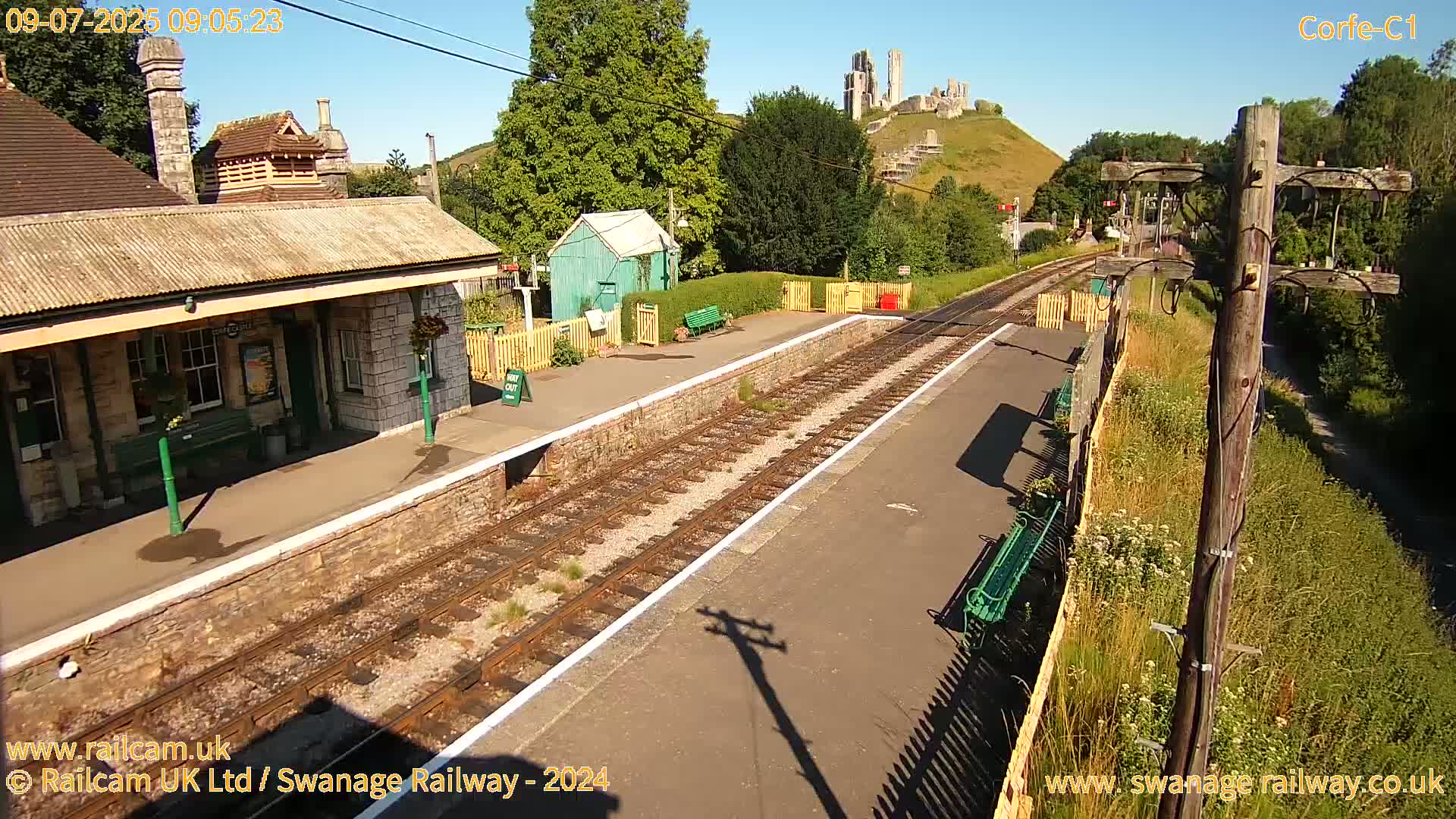 A train station platform with a stone building and a distant view of a castle on a hill under sunny skies.