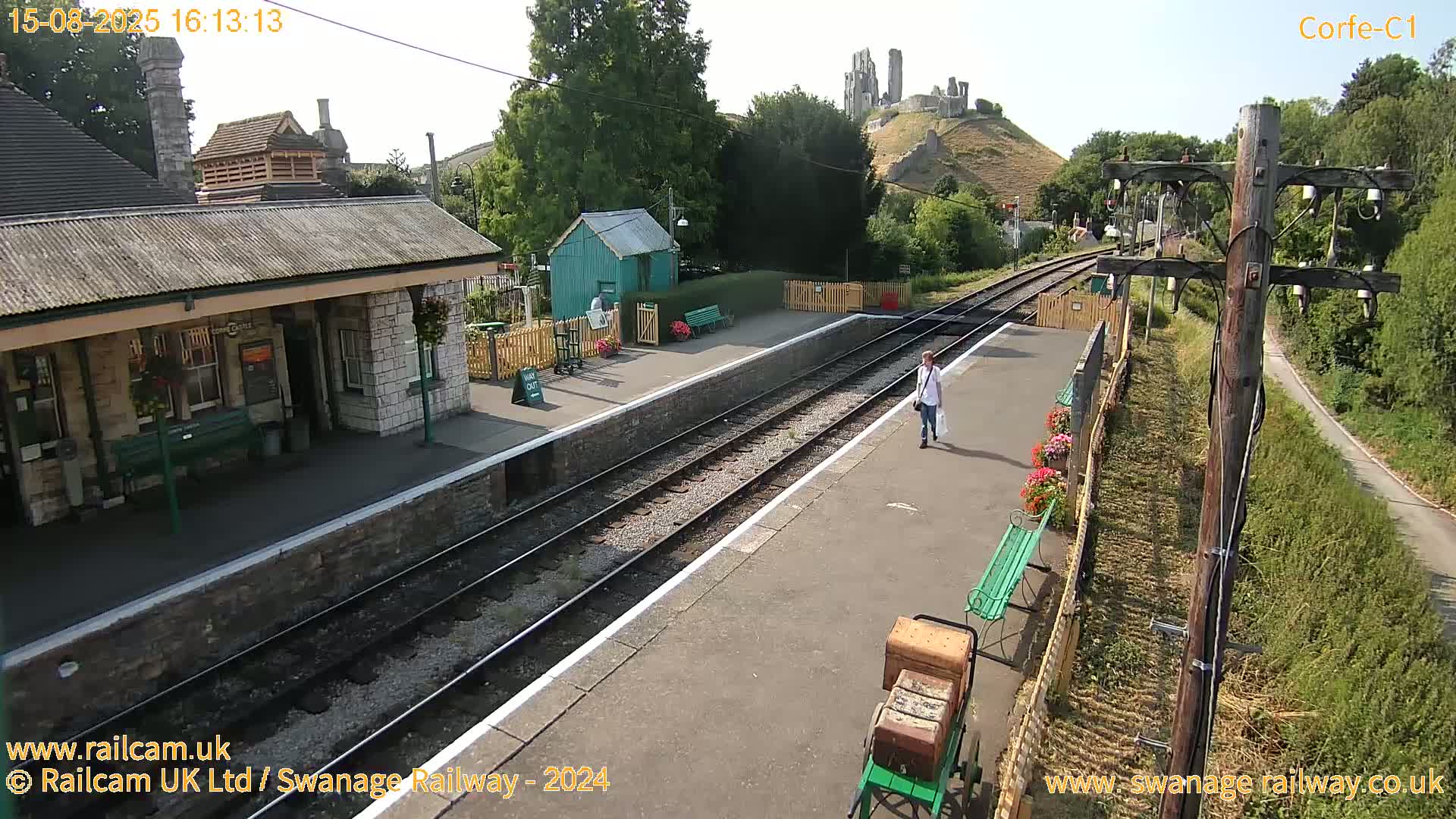 A train station platform with a stone building and a distant view of a castle on a hill under sunny skies.