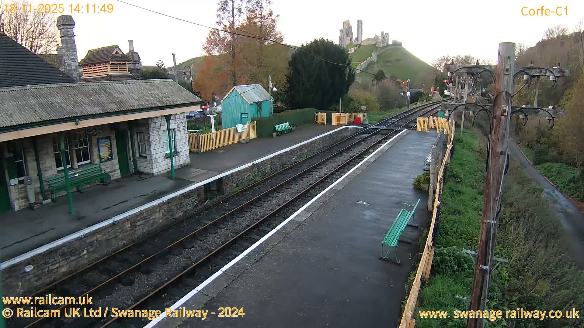 A train station platform with a stone building and a distant view of a castle on a hill under sunny skies.