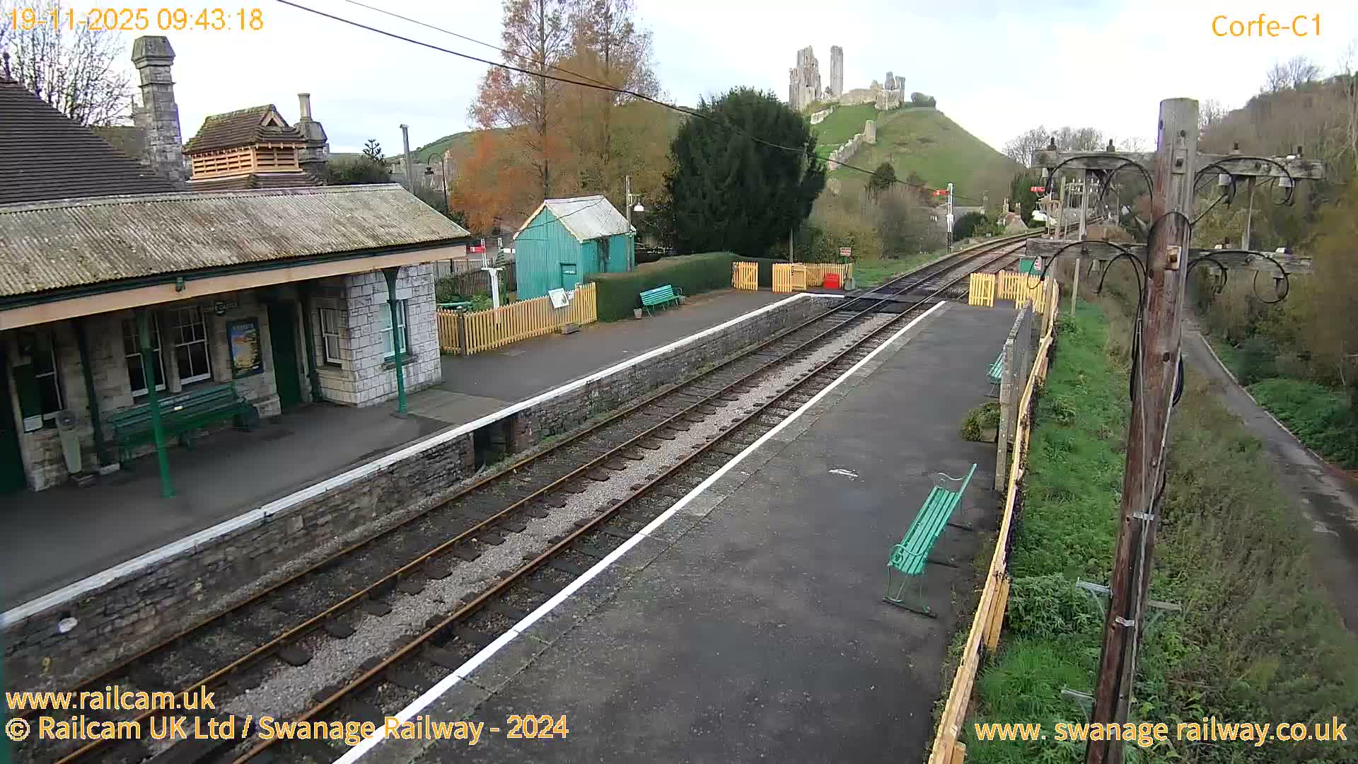 An overcast day illuminates a quiet train station platform with tracks stretching towards a prominent ruined castle perched atop a distant green hill.