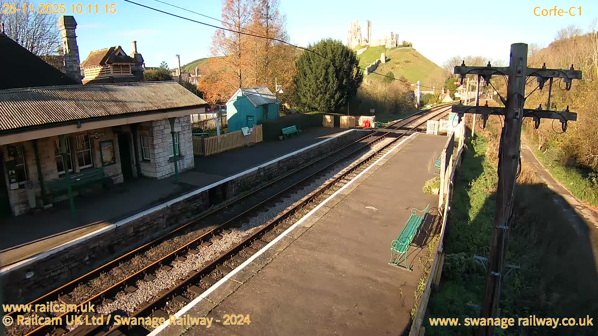 The image depicts a sunny view of a small train station with two platforms, tracks, and a stone building, framed by hills with autumn trees and distant castle ruins.