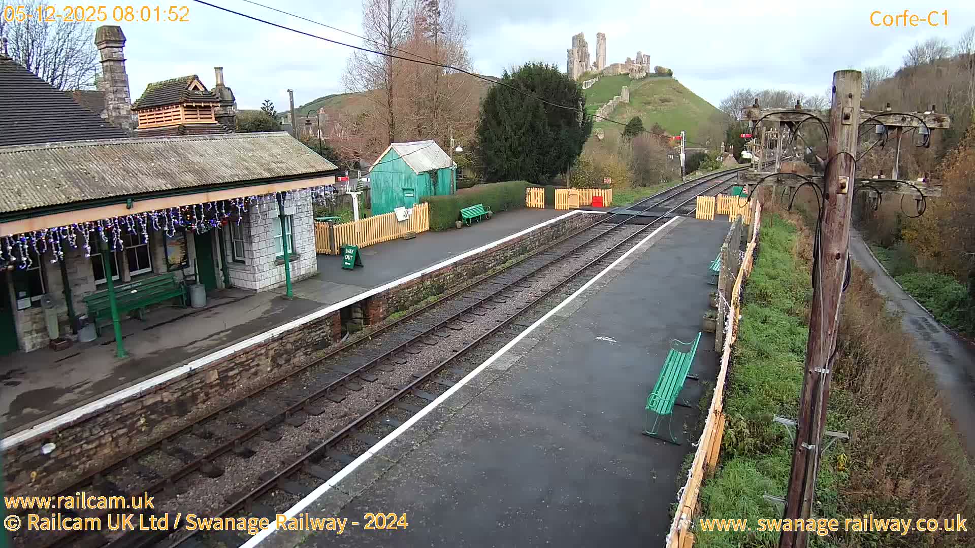 A historic train station building adorned with festive lights stands beside two empty tracks, facing a green hill topped by the ruins of Corfe Castle, all under an overcast sky.