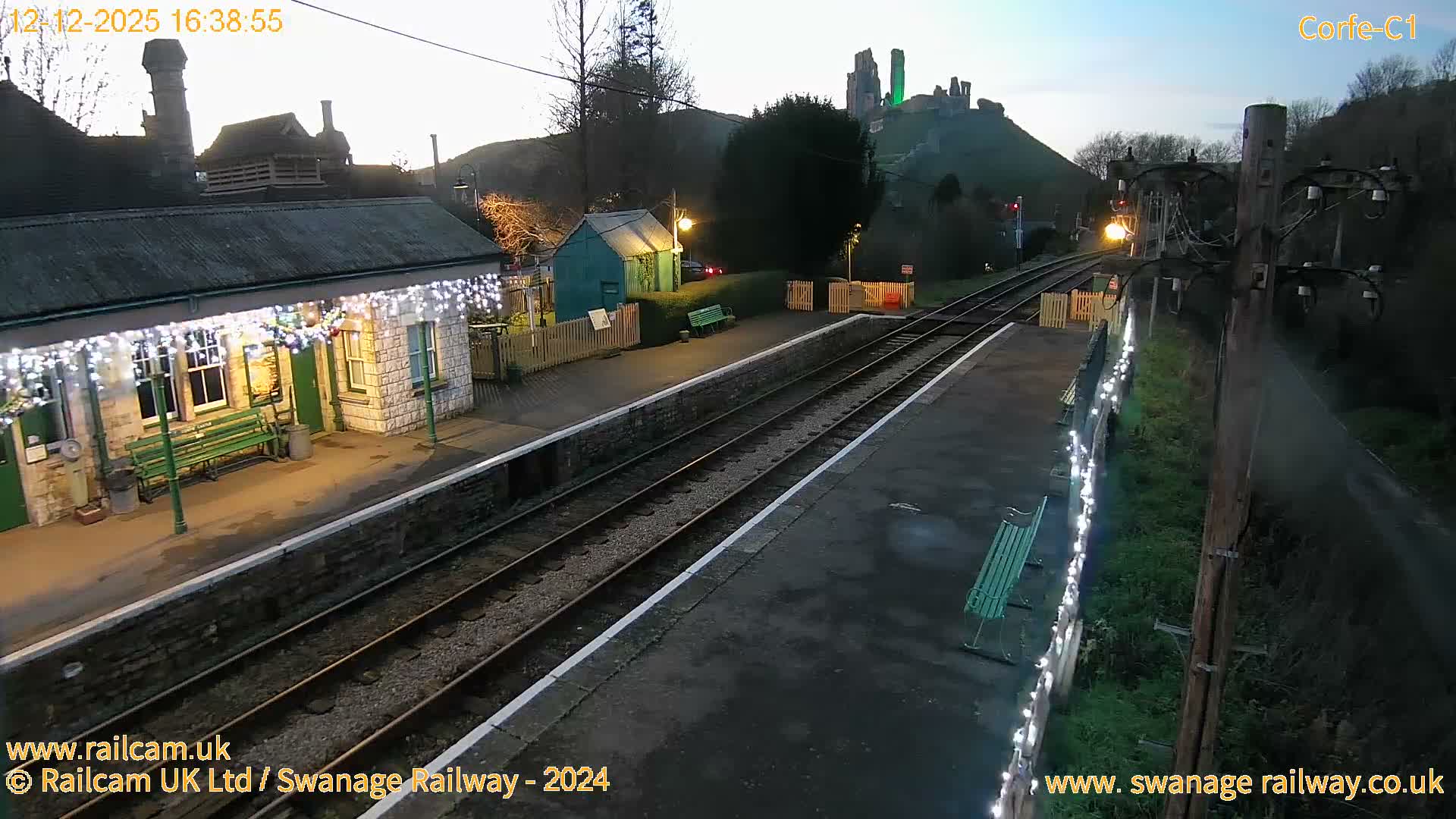 An outdoor train station is adorned with festive white lights under a clear, dusky sky, as a train's headlight shines on the tracks and Corfe Castle glows green on a distant hill.