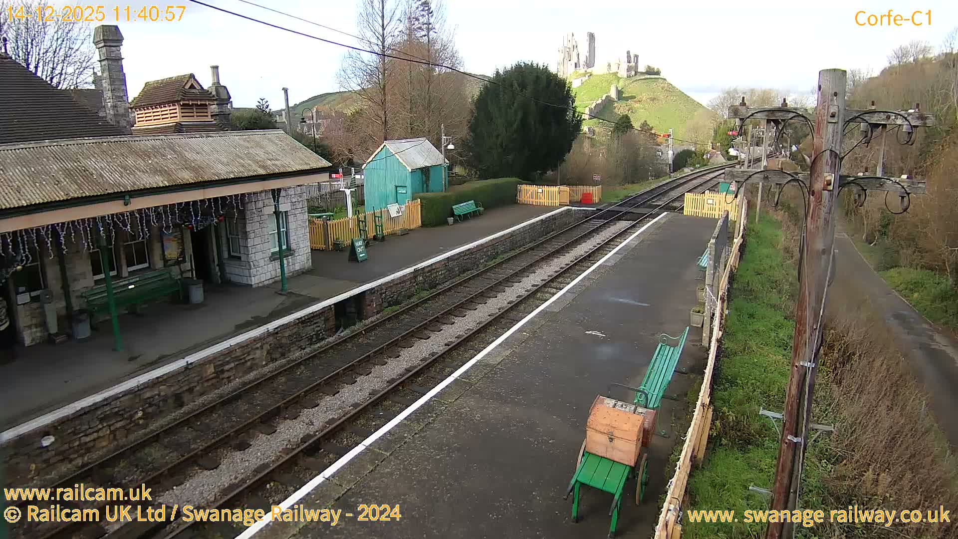 An outdoor train station is adorned with festive white lights under a clear, dusky sky, as a train's headlight shines on the tracks and Corfe Castle glows green on a distant hill.