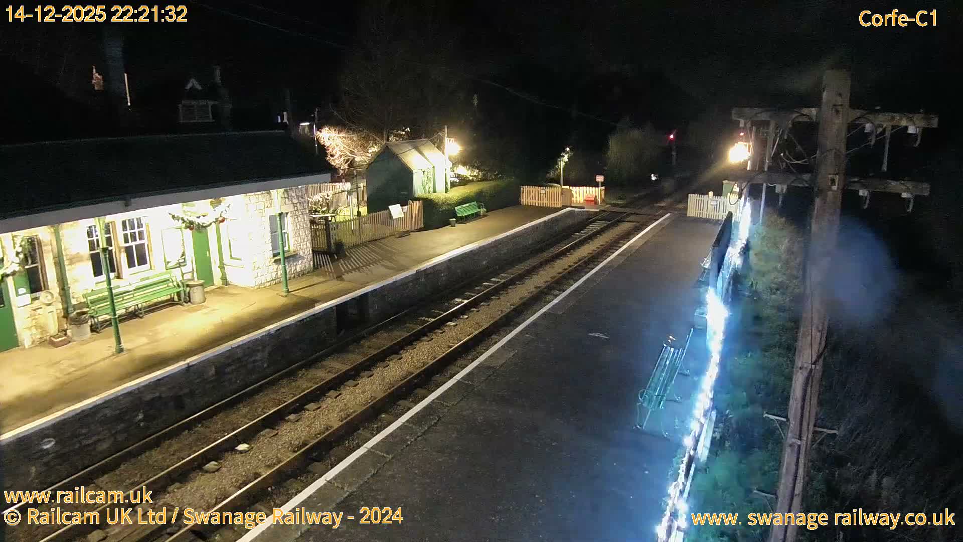 An outdoor train station is adorned with festive white lights under a clear, dusky sky, as a train's headlight shines on the tracks and Corfe Castle glows green on a distant hill.