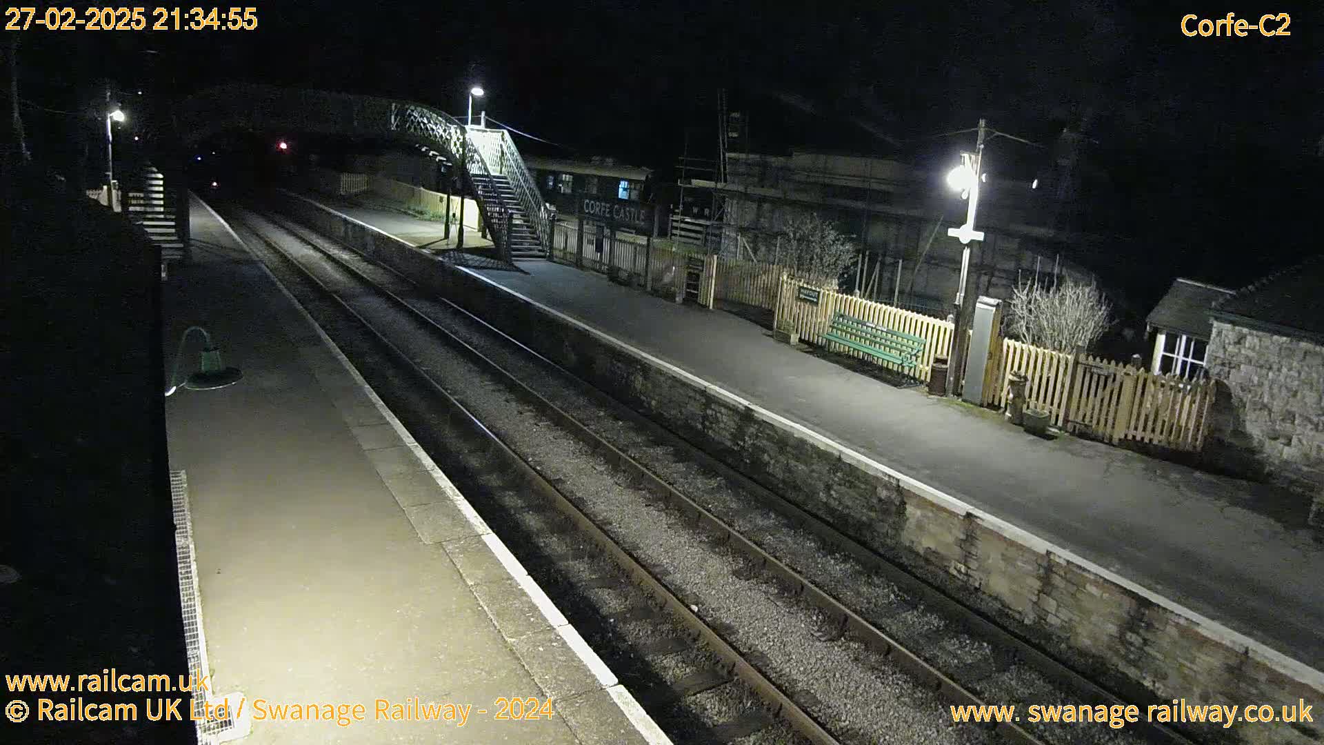 A nighttime view of a deserted train station platform with two sets of train tracks, a pedestrian bridge, and several buildings.