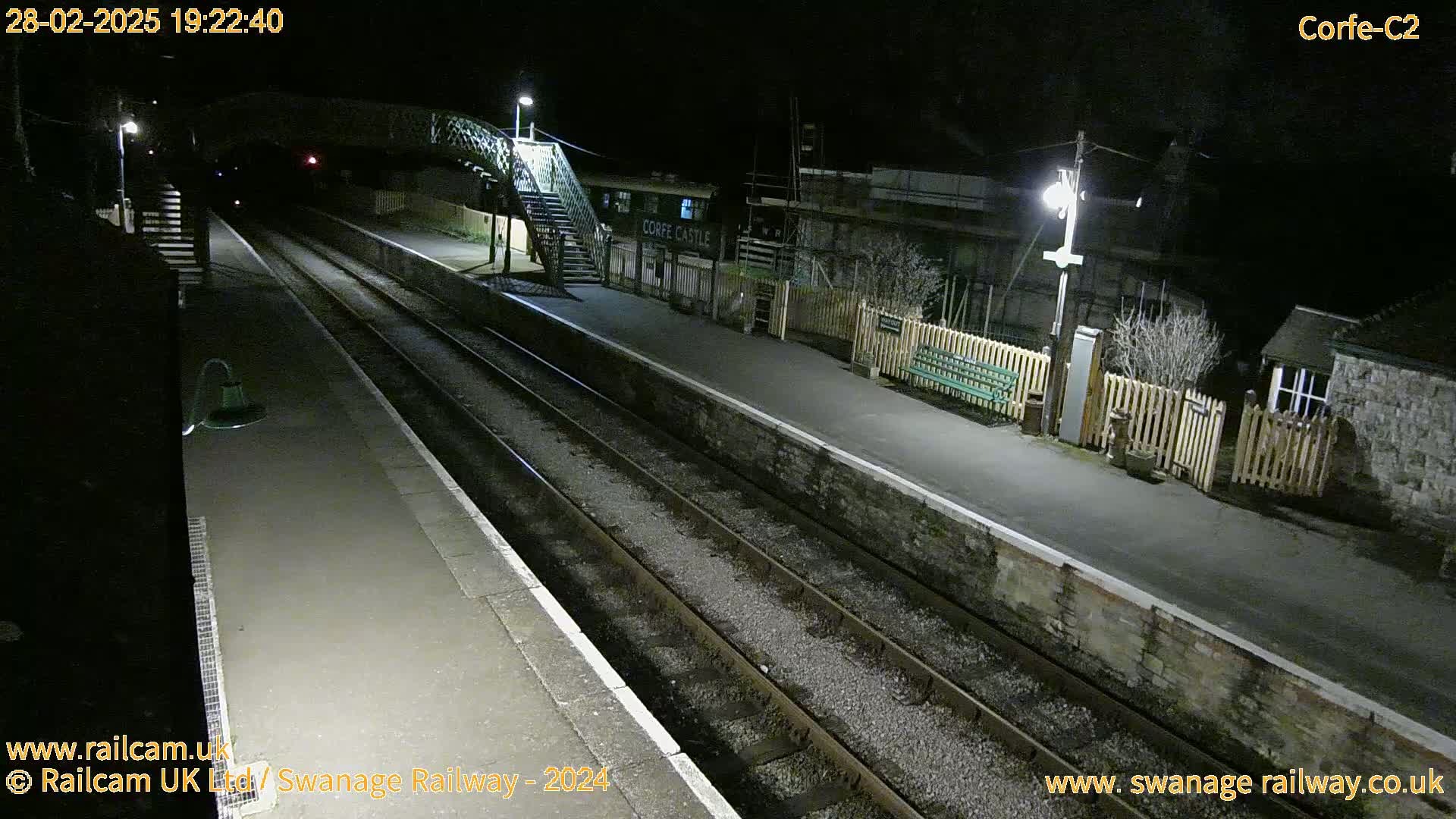 A nighttime view of a deserted train station platform with two sets of train tracks, a footbridge, and some buildings.