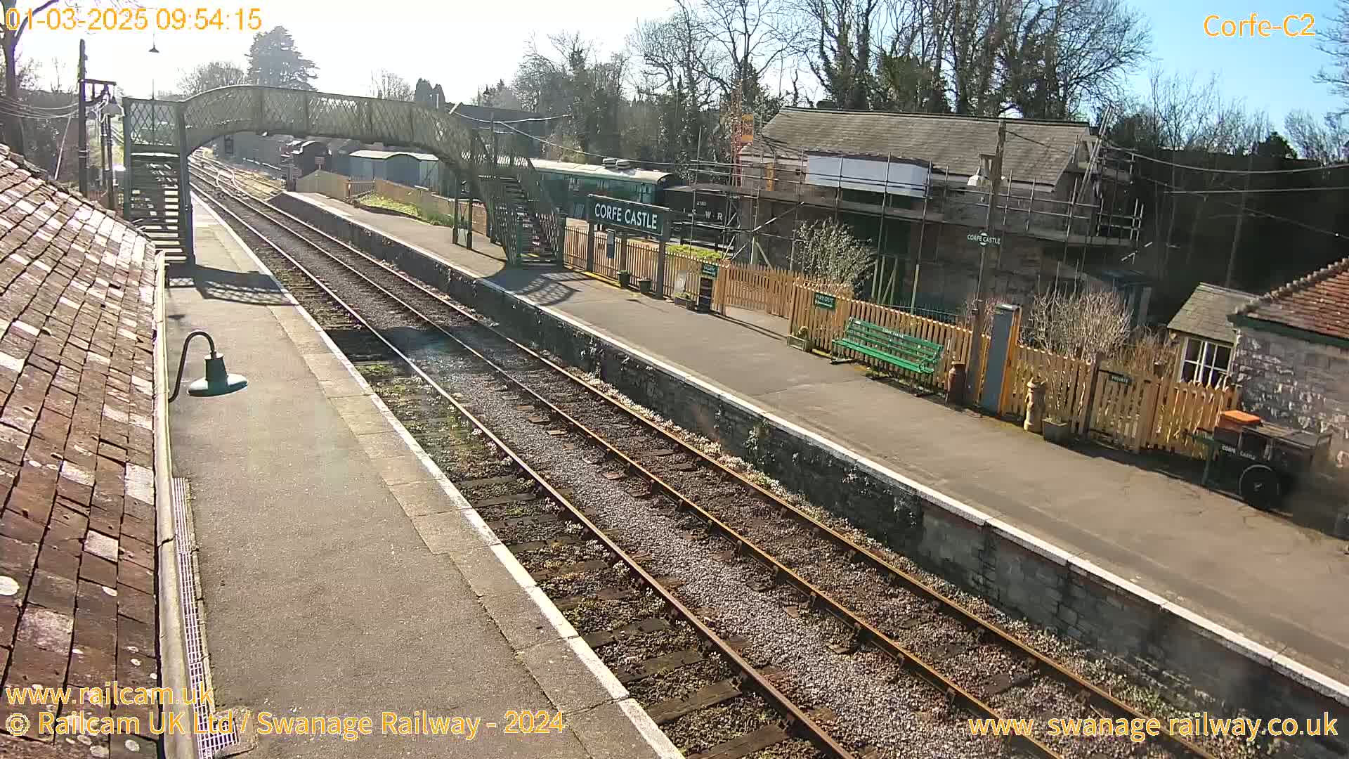 A sunny day at a train station shows two parallel train tracks, a train car, and several station buildings, one of which is under construction.