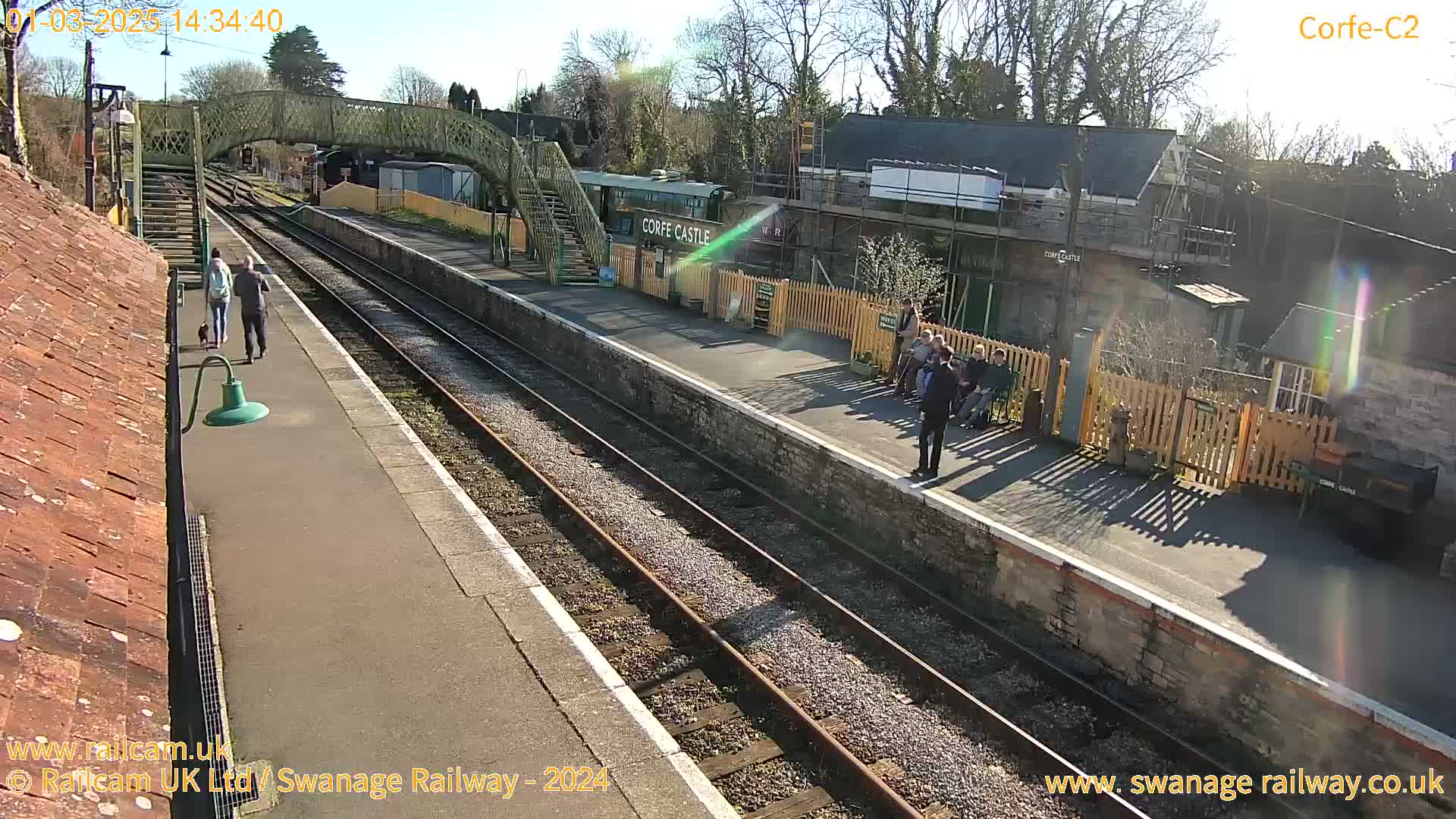 A sunny day at a train station shows a couple walking a dog on the platform, several people waiting near a building under construction, and train tracks extending into the distance.