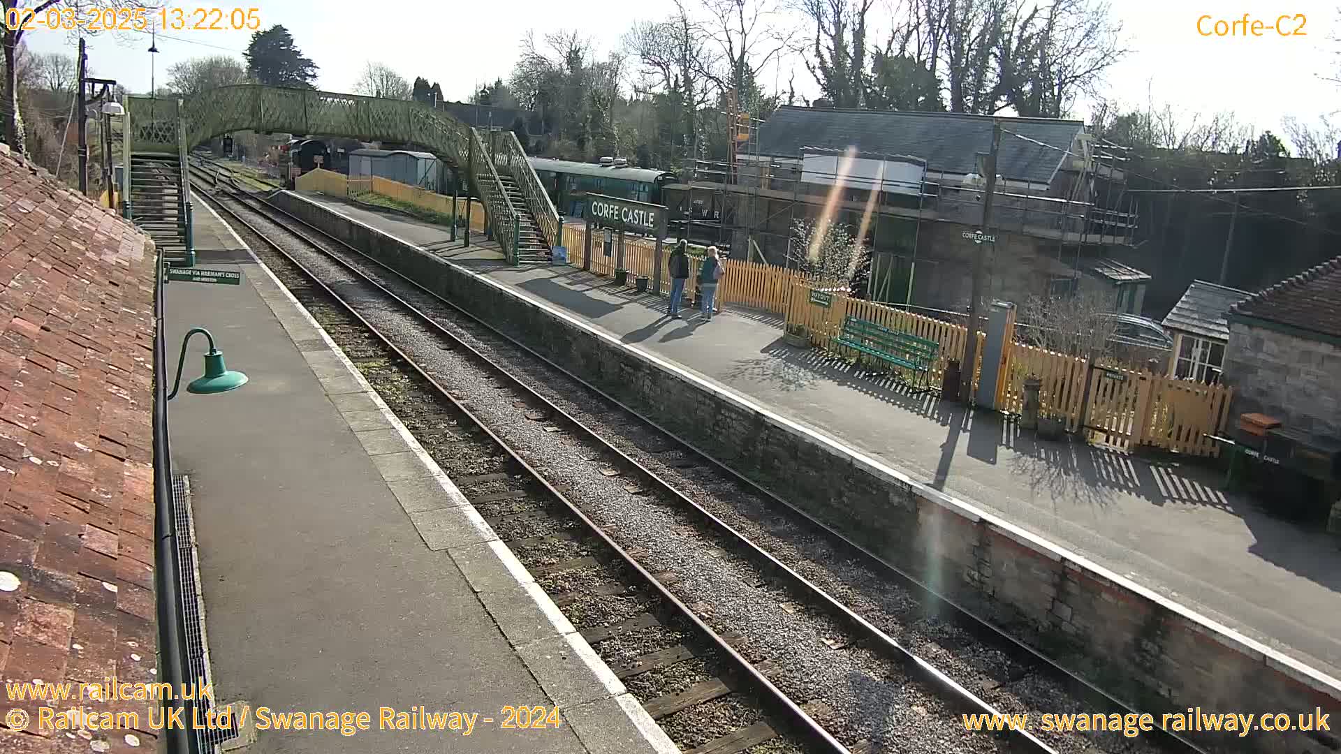 A sunny view of a train station with two parallel train tracks, a platform, a pedestrian bridge, and a few people standing near a building under construction.