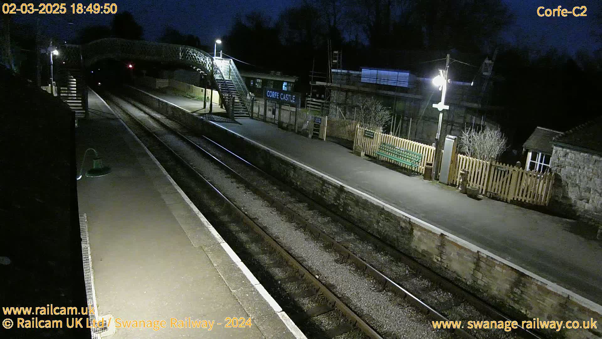 A nighttime view of a deserted train station platform with two train tracks, a footbridge, and a small building.