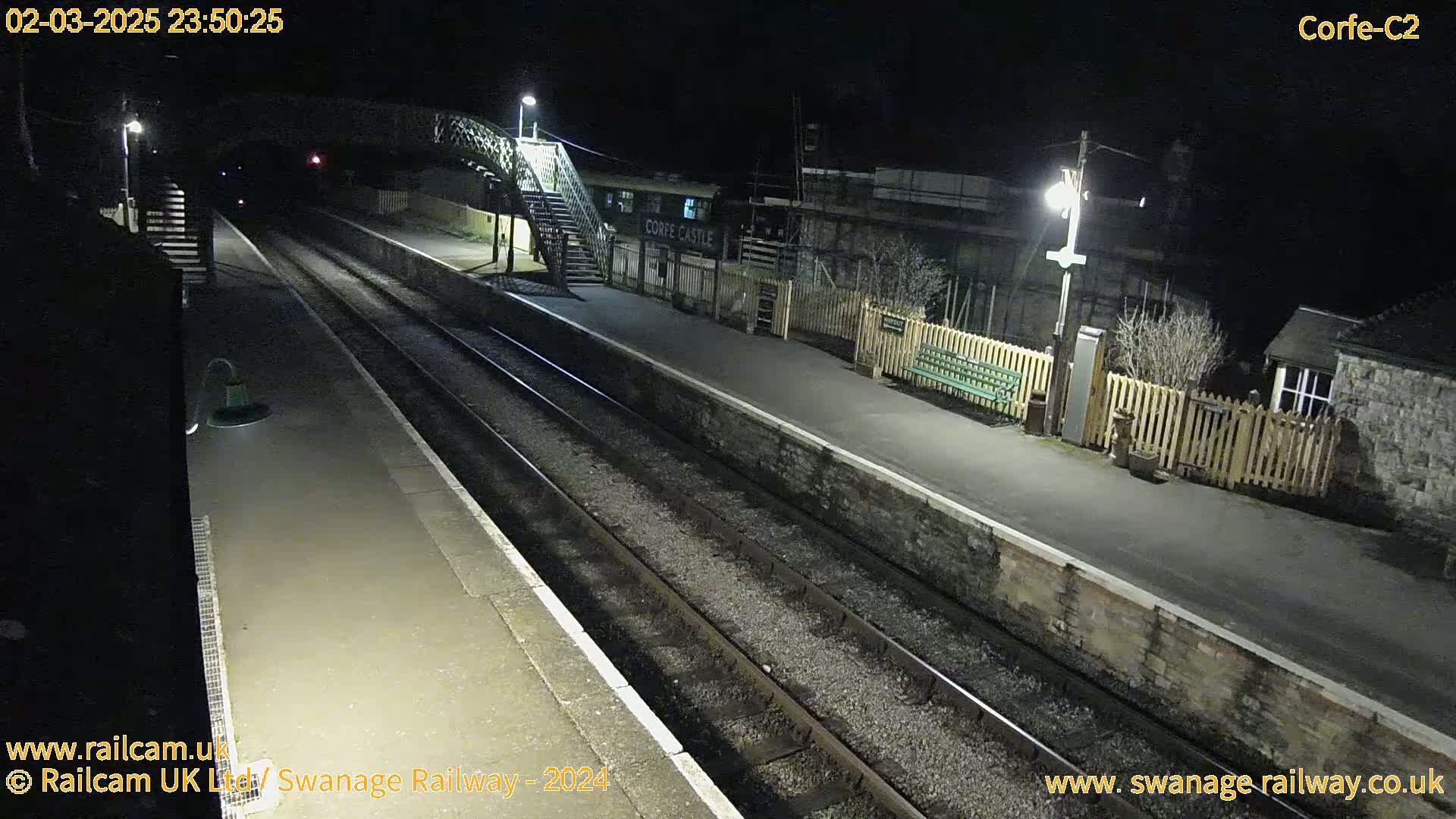 A nighttime view of a deserted train station platform with two sets of train tracks, lit by several lamps.