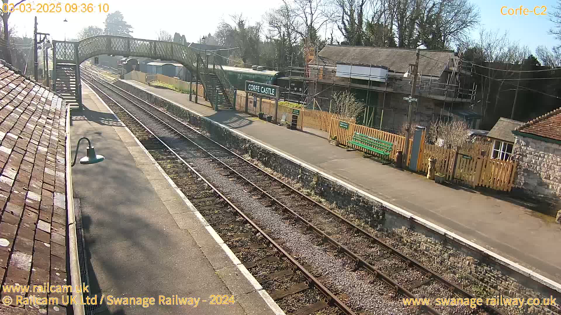 A sunny view of a train station with a railway line, platform, footbridge, and several buildings, one of which is under construction.
