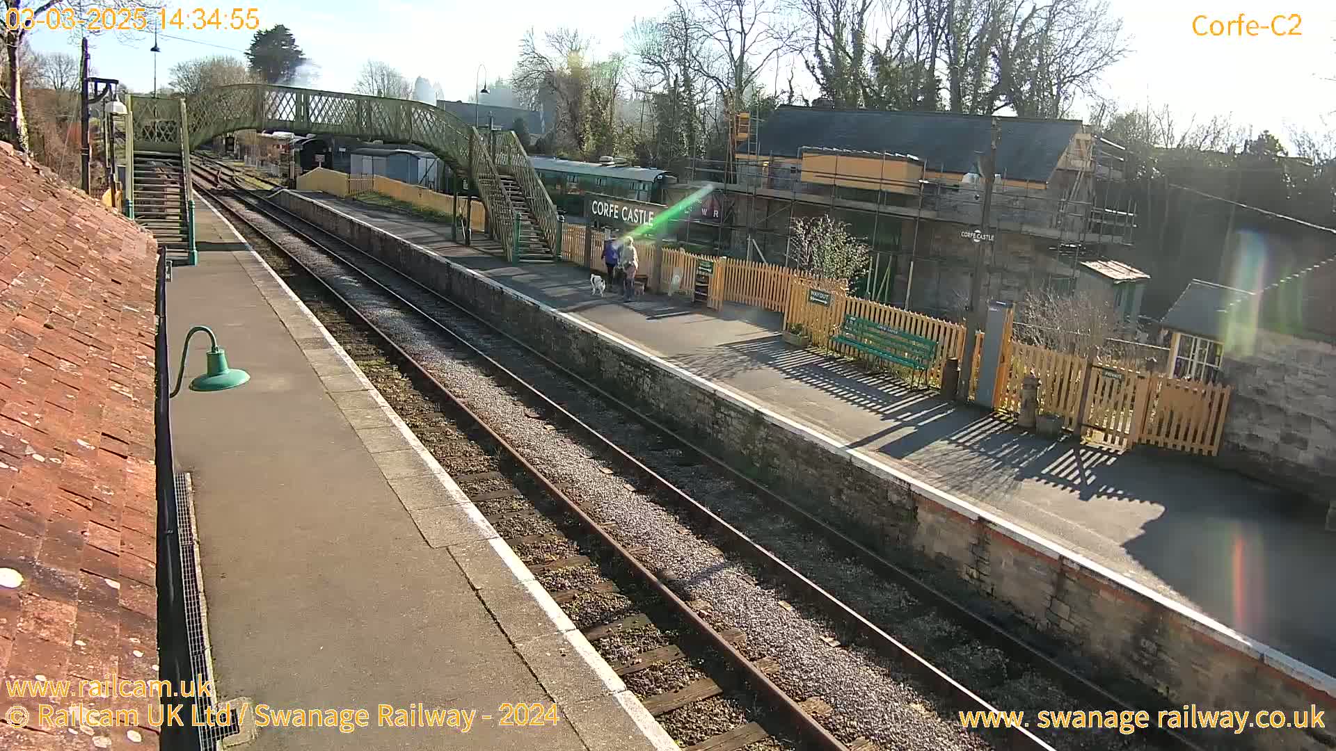 A sunny view of a train station platform with two people walking dogs, railway tracks, and buildings.