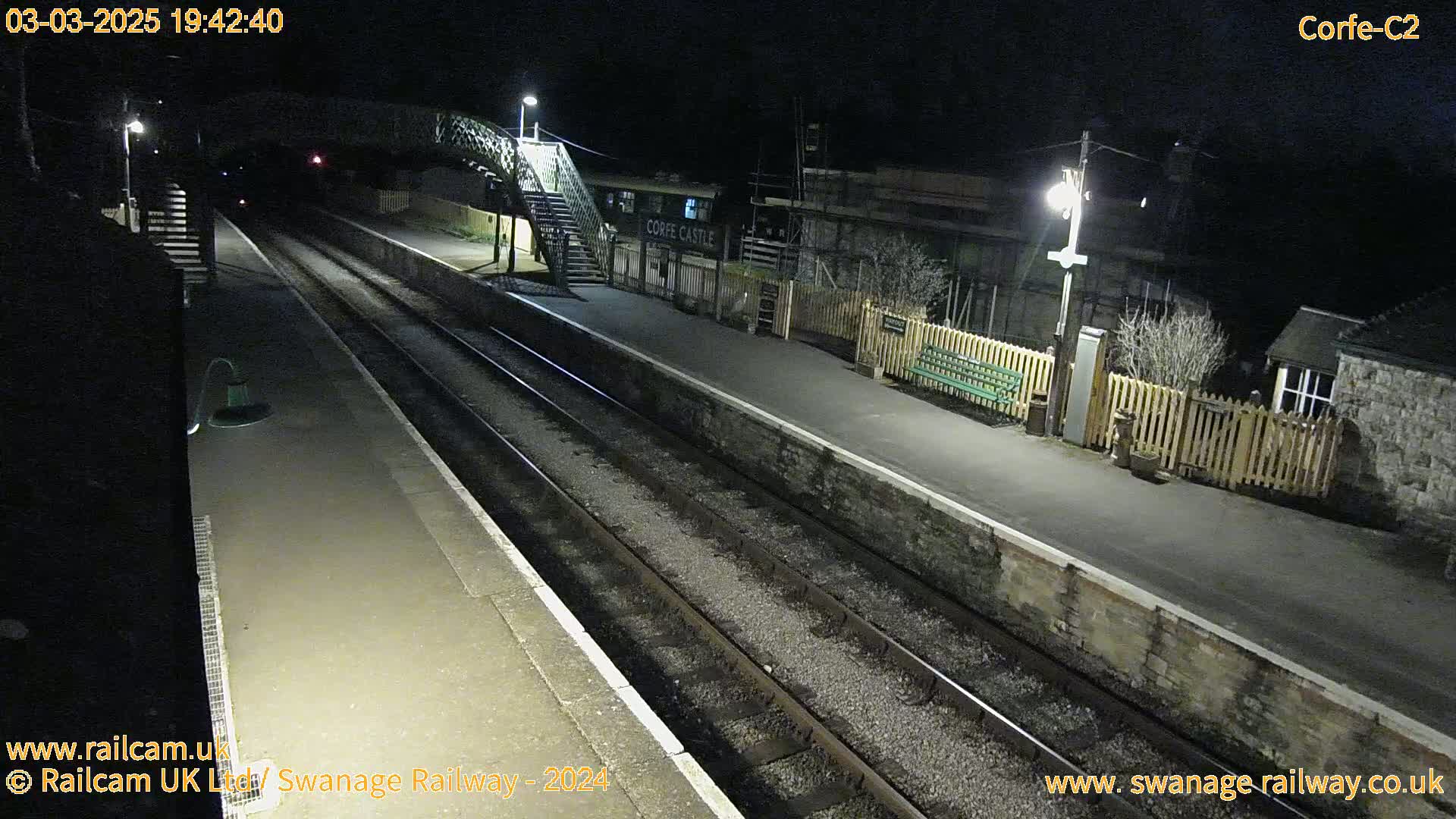 A nighttime view of a deserted train station platform with two sets of train tracks, lit by overhead lamps.
