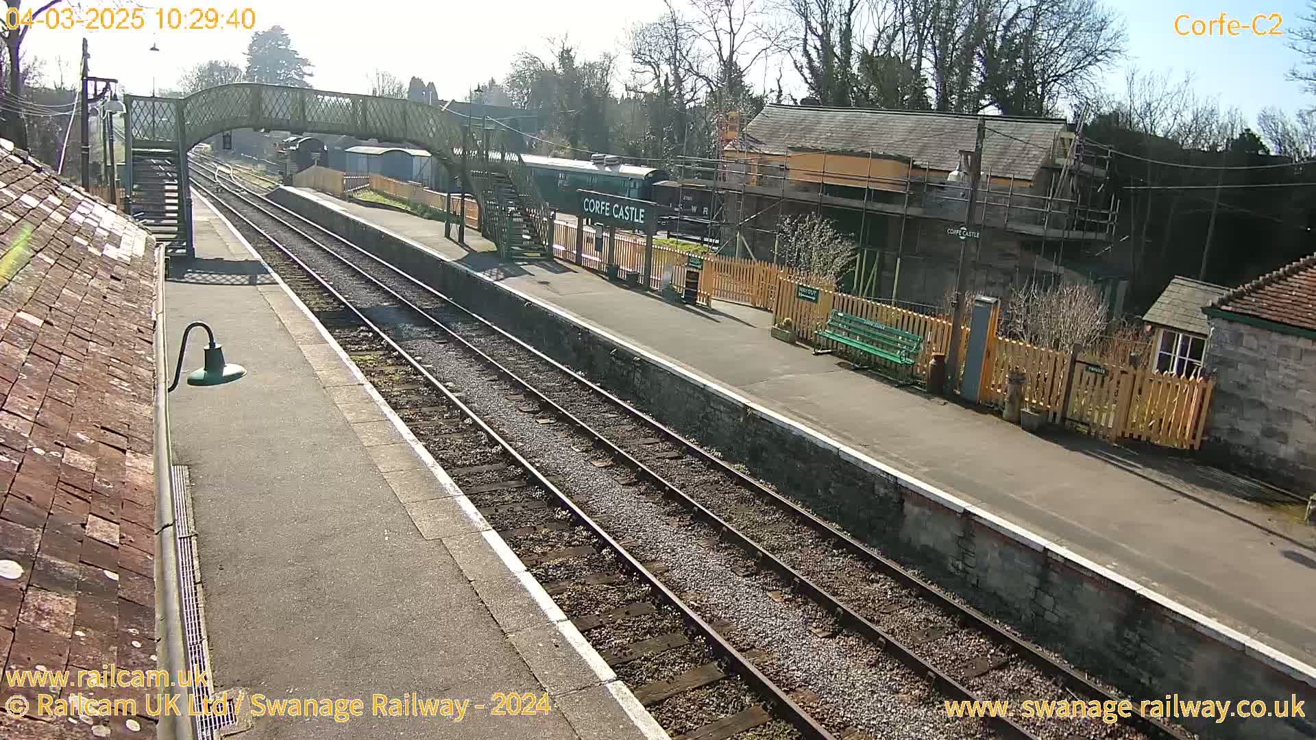 A sunny view of a train station with multiple tracks, a train at the platform, and a footbridge overhead.