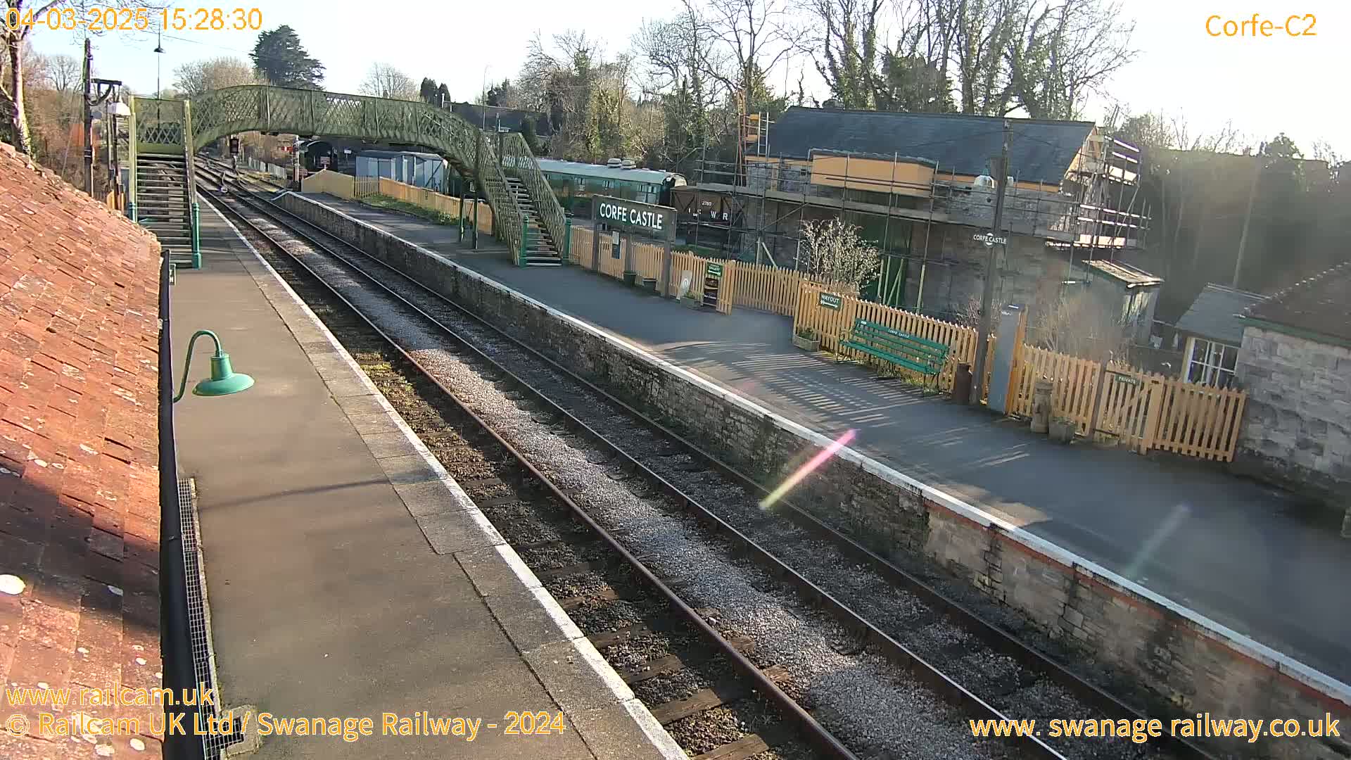 A sunny train station with two parallel train tracks, a railway carriage, a footbridge, and several buildings, one of which is under construction.