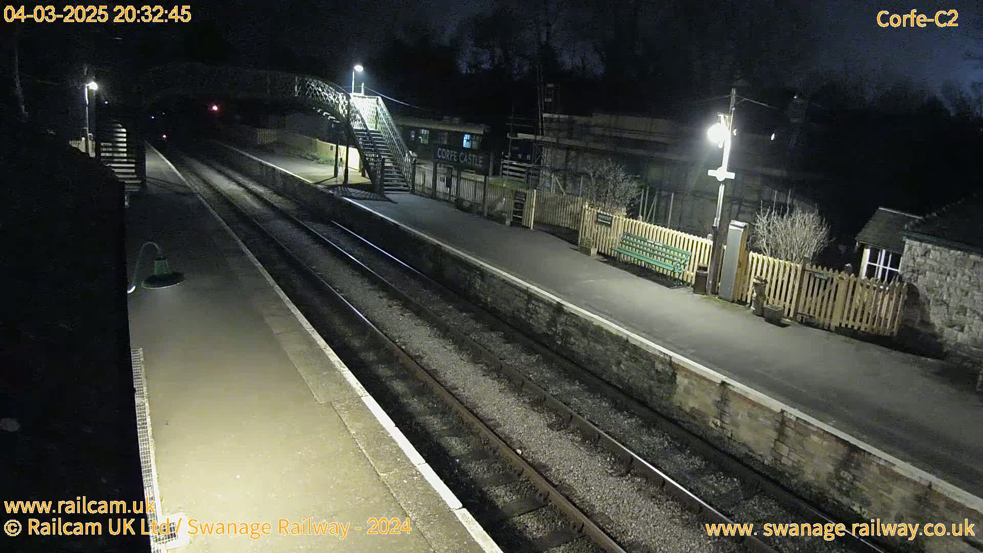 A nighttime view of a deserted train station with two train tracks, a platform, and a footbridge under artificial lighting.