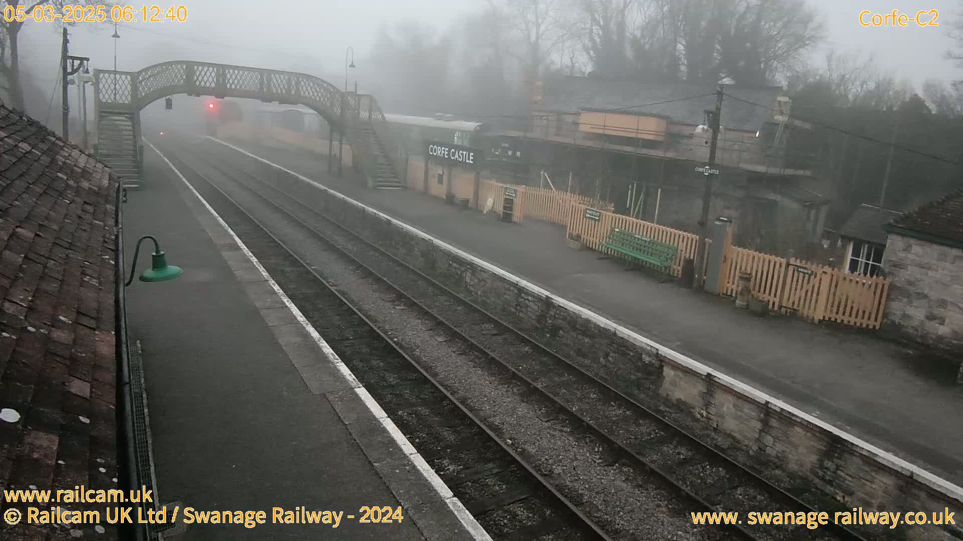 A foggy view of a train station with two tracks, a pedestrian bridge, and a station building.