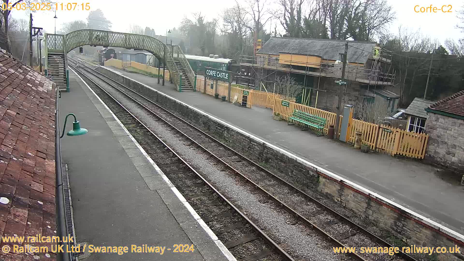 A train station on an overcast day features a railway platform, tracks, a pedestrian bridge, and several buildings, one of which is under construction.