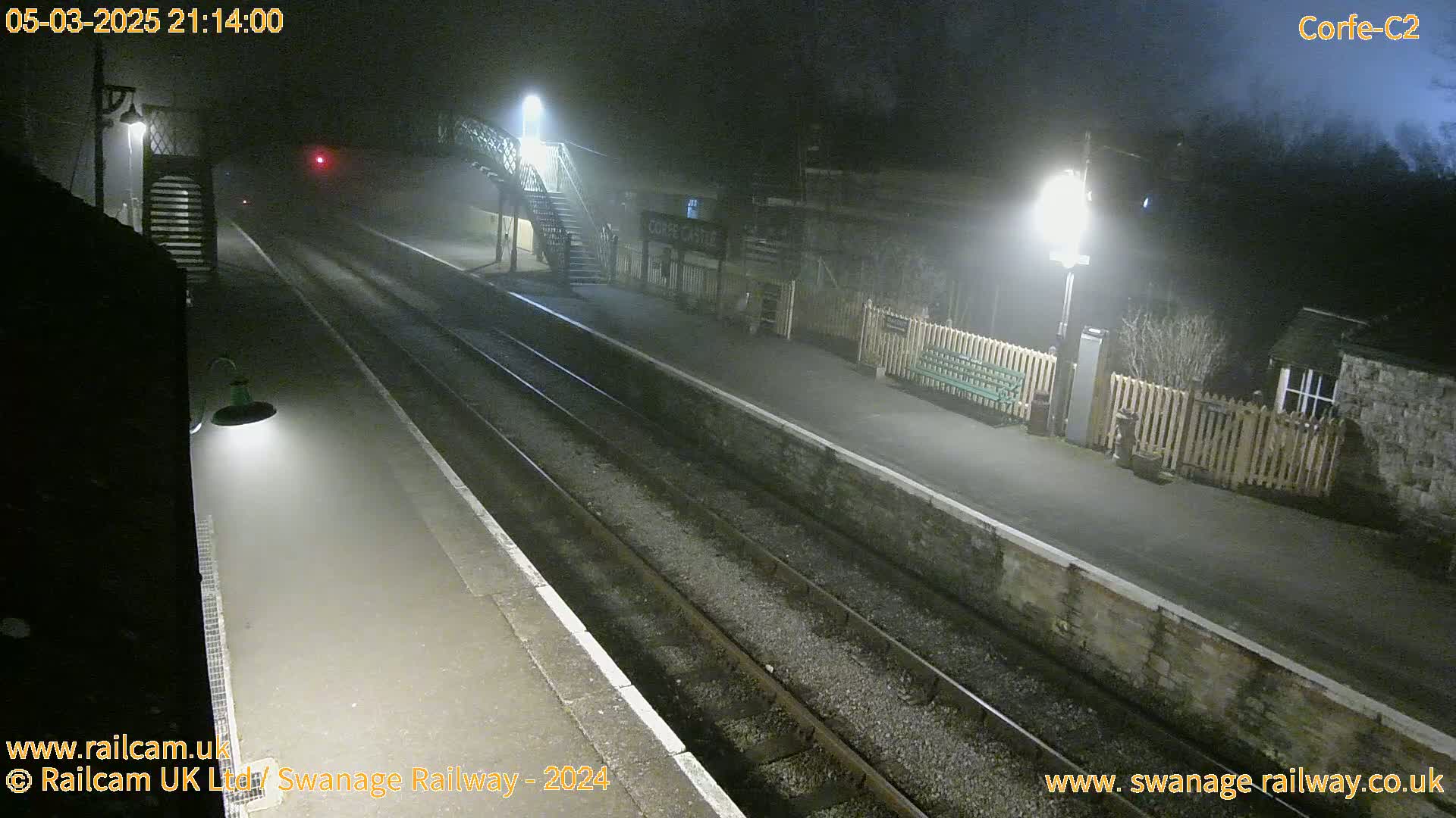 A foggy night scene at a train station with two sets of train tracks, platforms, and lights illuminating the area.