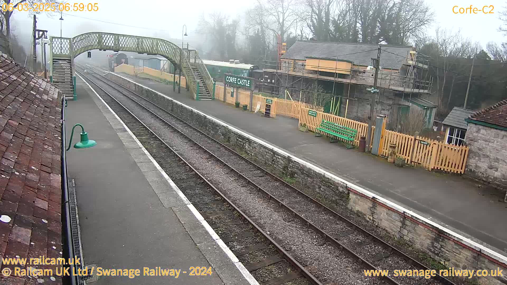 A train station on an overcast day, featuring a railway platform, tracks, pedestrian bridge, and several buildings.