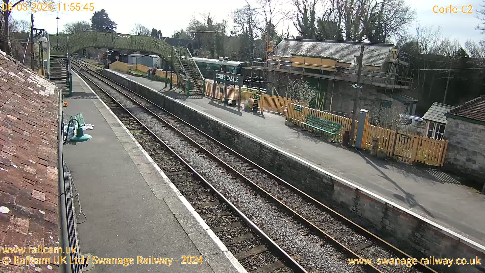 A sunny day at a train station with a passenger train and a footbridge, showing a partially-constructed building.