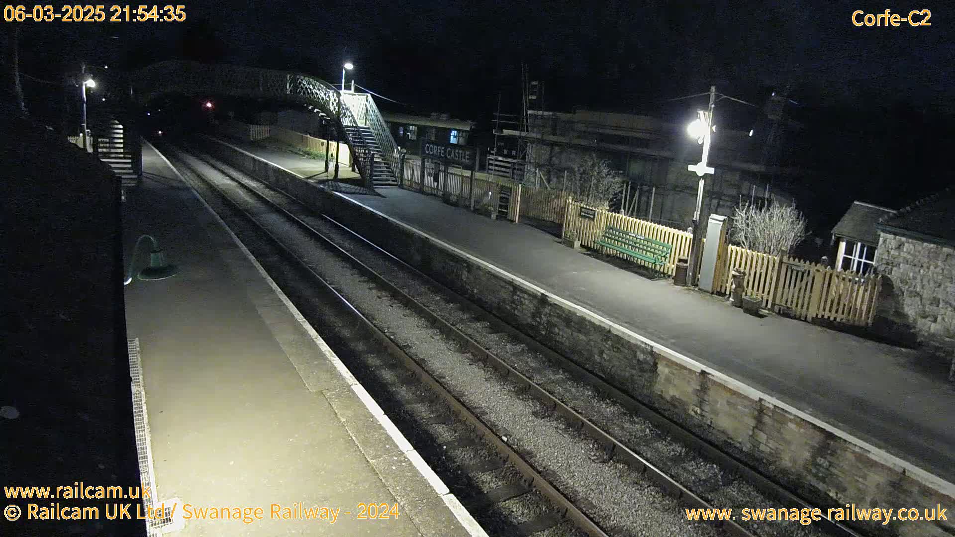 A nighttime view of a deserted train station platform with two train tracks, a footbridge, and a small building.